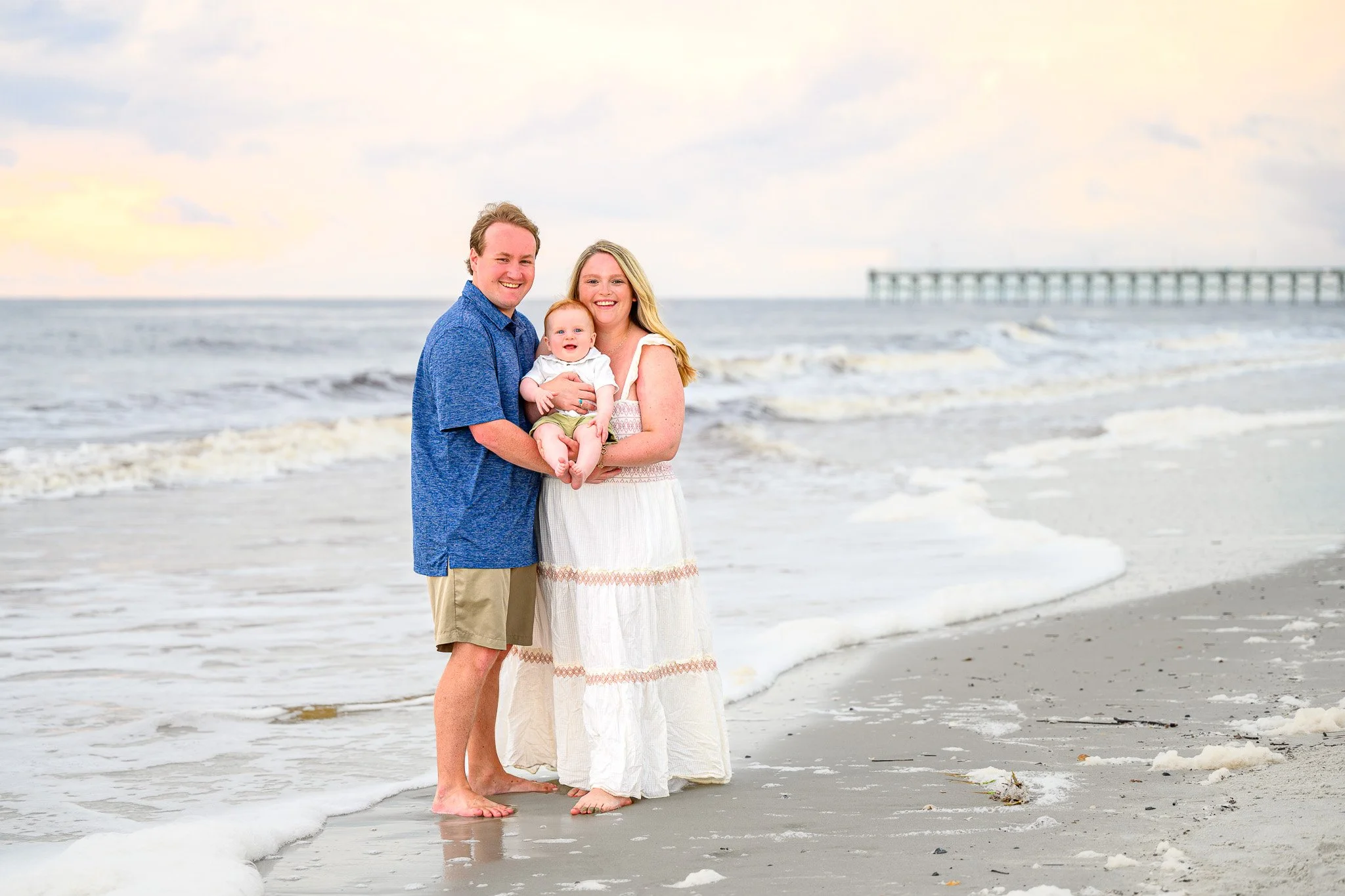 A smiling family of three, a man, woman, and baby, standing barefoot on a beach with waves and a pier in the background during sunset at Oak Island, NC