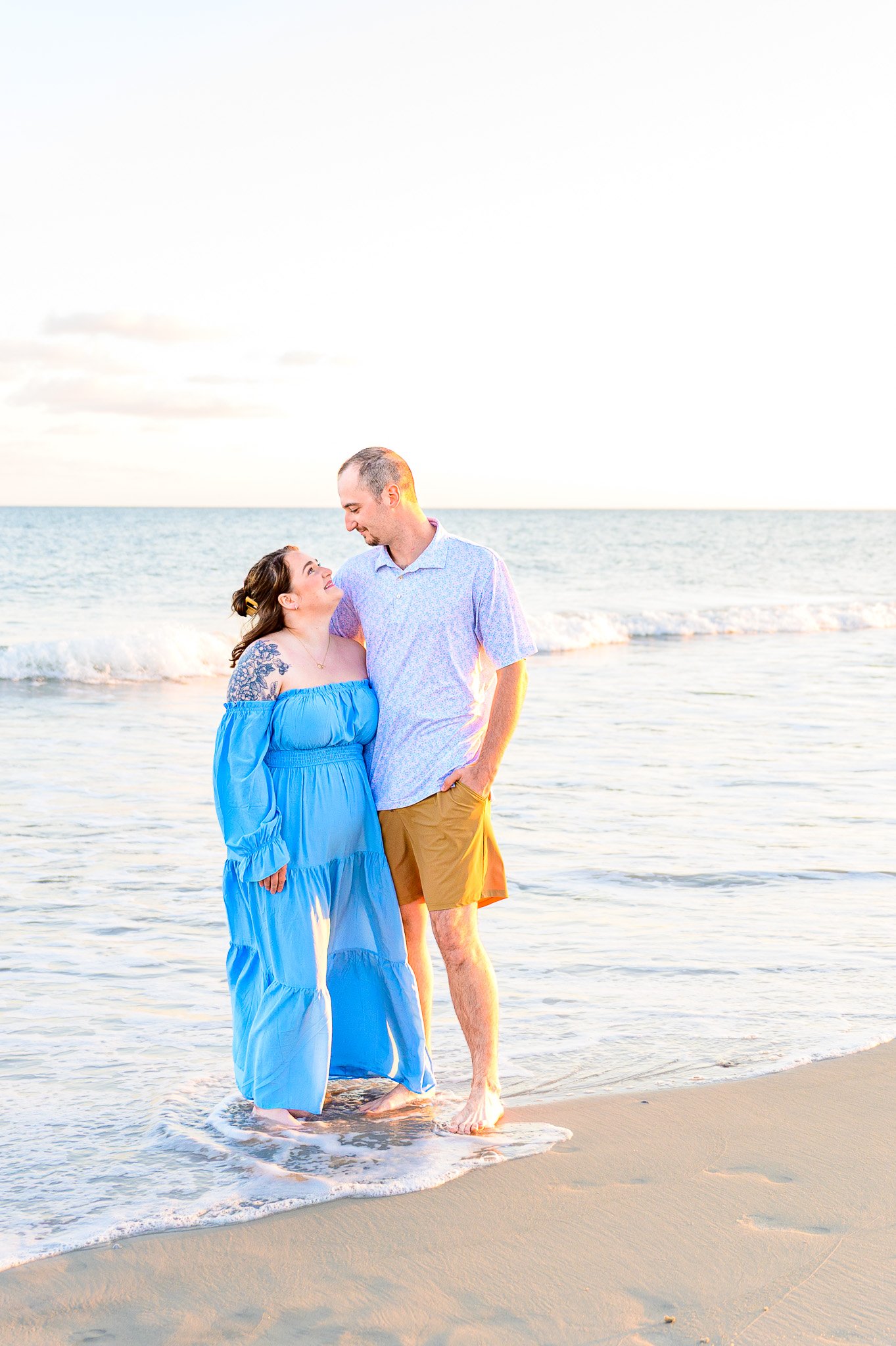 Family beach photo session on Oak Island, NC with a couple standing barefoot in the ocean during golden hour sunset, captured by Oak Island family photographer AEM Creative Photography.
