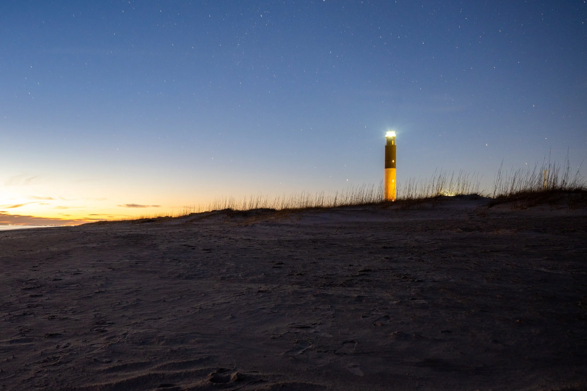 Oak Island Lighthouse illuminated at dusk with stars appearing above the beach and dunes in North Carolina.