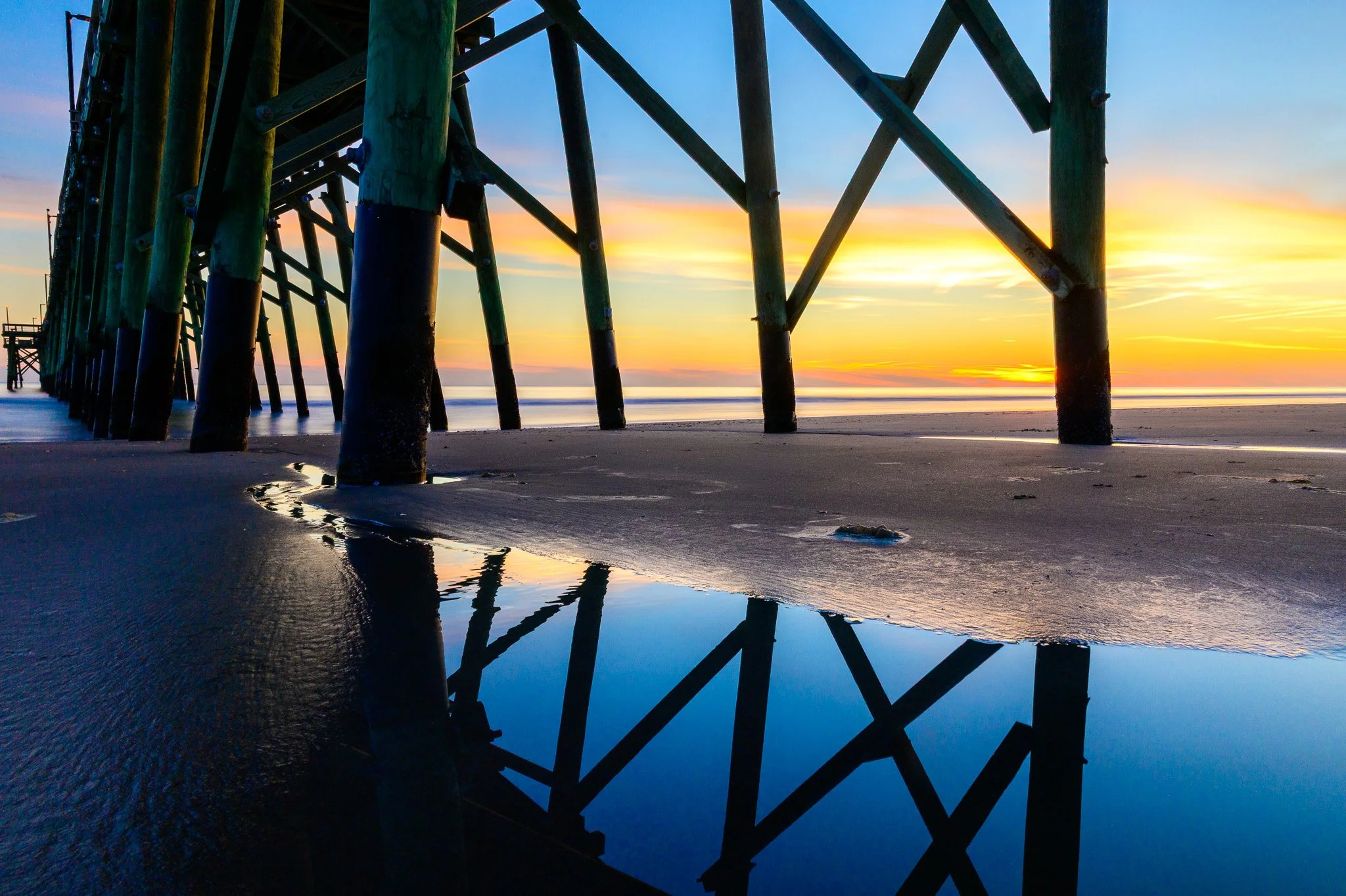 Sunrise reflections beneath Oak Island Pier with colorful skies and wet sand on Oak Island, North Carolina.