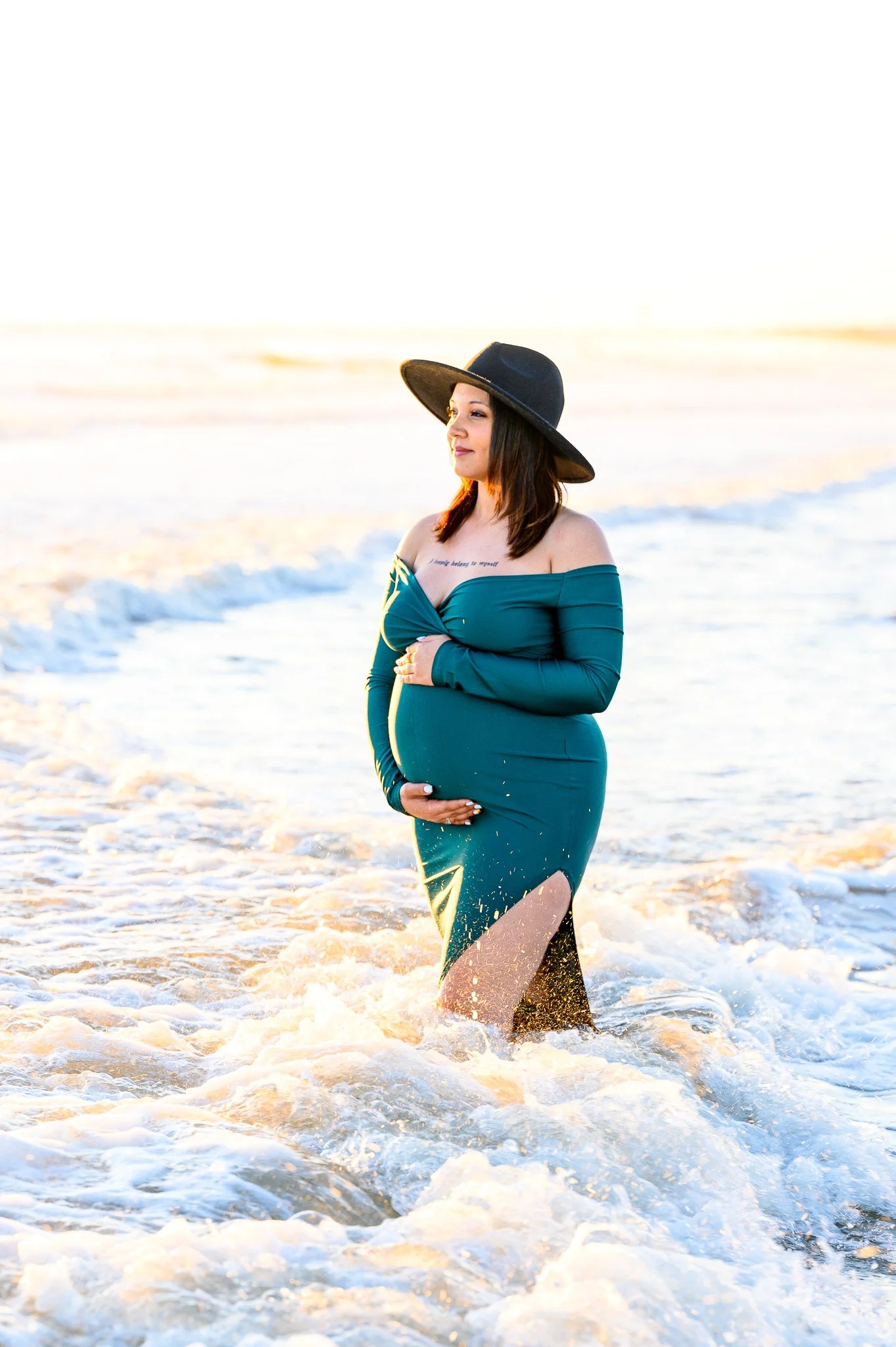 A pregnant woman in a teal dress and wide-brimmed black hat standing in the surf at the beach during sunset at Oak Island, NC.