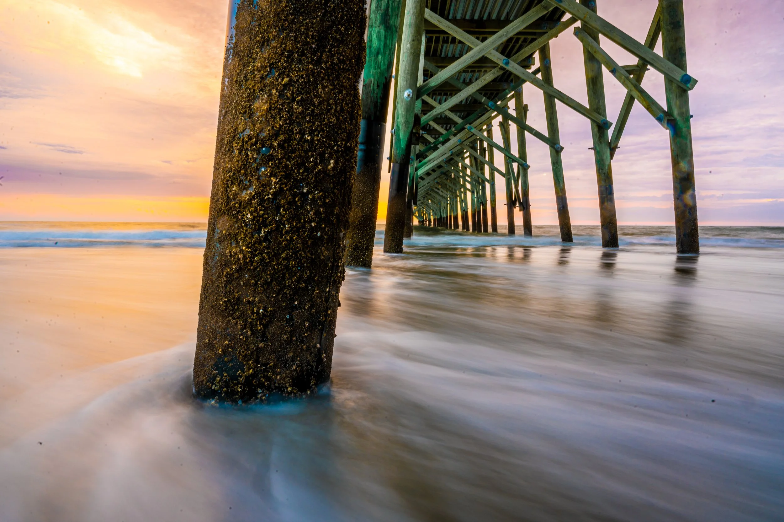 Sunset view beneath the Oak Island Pier with waves washing across the beach in Oak Island, North Carolina.