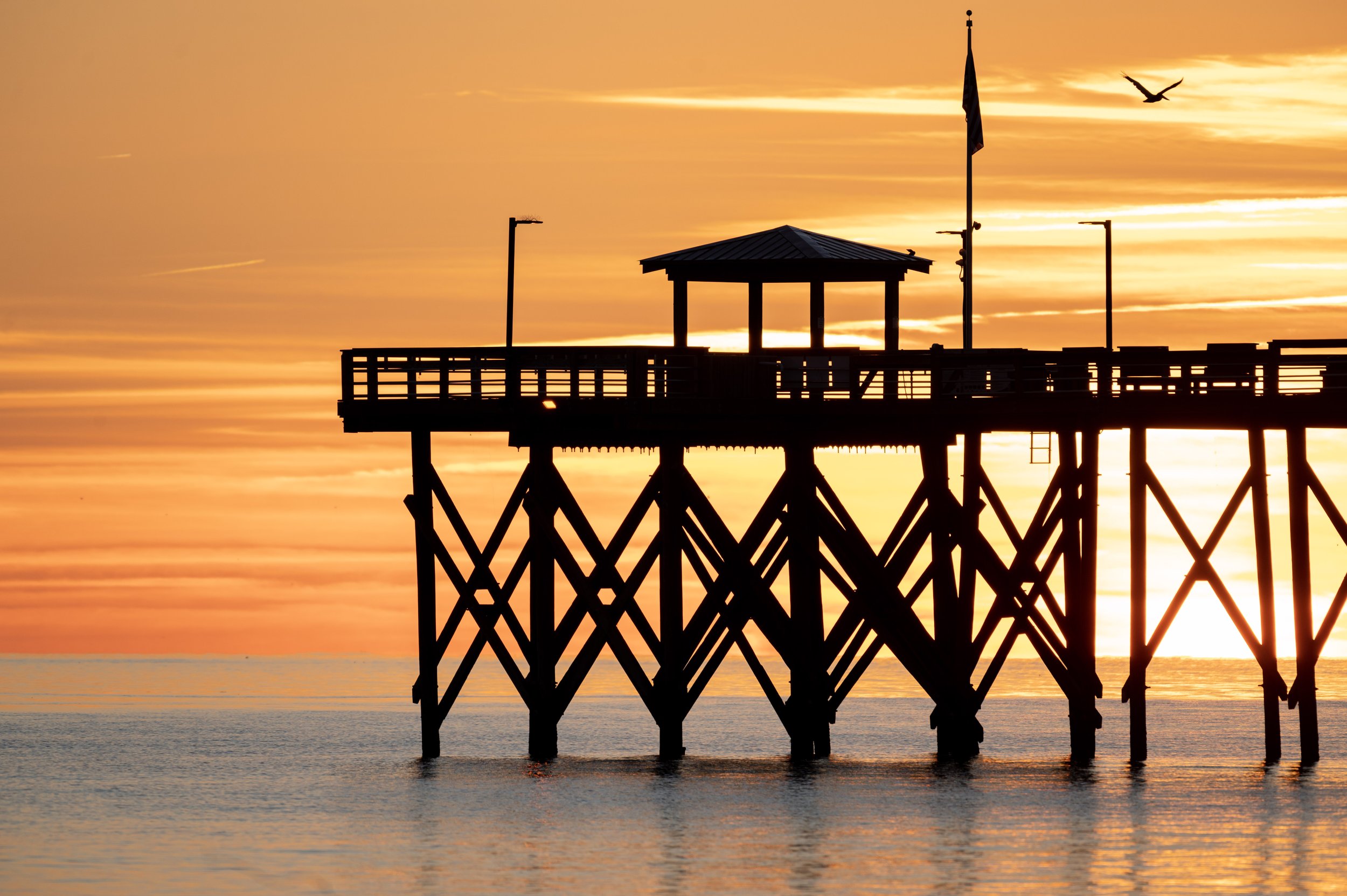 Silhouette of Oak Island Pier at sunrise with colorful orange skies and calm ocean waters in Oak Island, North Carolina.