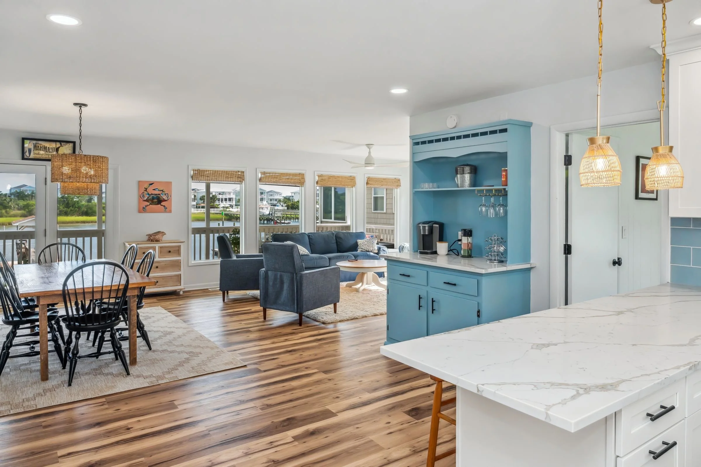 Open-concept living room and dining area with large windows overlooking water, blue and gray furniture, wooden dining table, and kitchen with white countertops and blue cabinets at Oak Island, NC.
