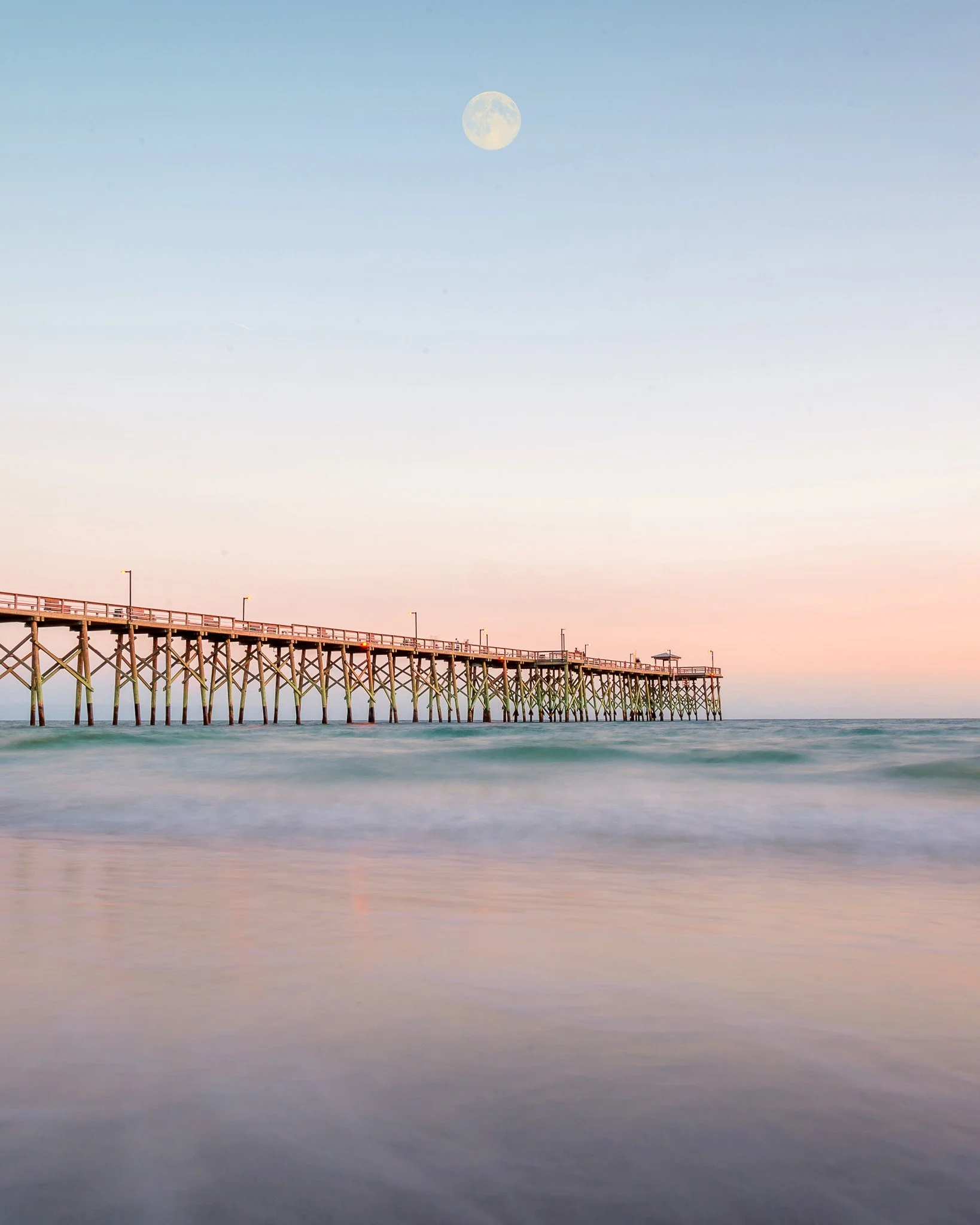 Oak Island Pier extending into the Atlantic Ocean during a calm sunrise with the moon visible above the water in Oak Island, North Carolina.