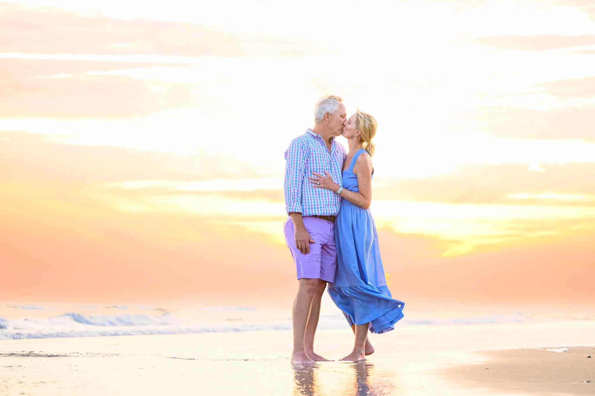 A senior couple sharing a kiss on the beach at sunset. The man is wearing a multicolored checkered shirt and purple shorts, and the woman is in a blue dress. They are standing barefoot on the sand near the water on Bald Head Island.