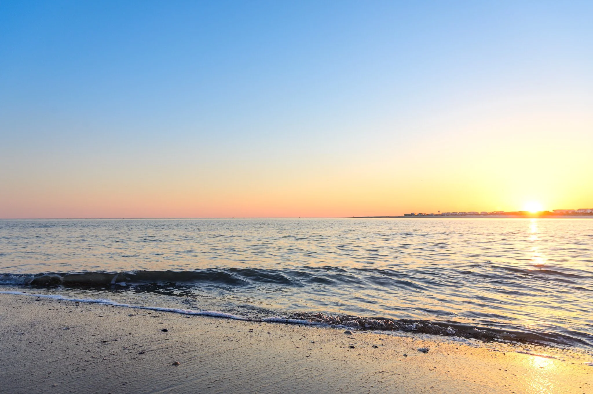 Sunset over the water at The Point on Oak Island, North Carolina, with gentle waves rolling onto the shoreline.