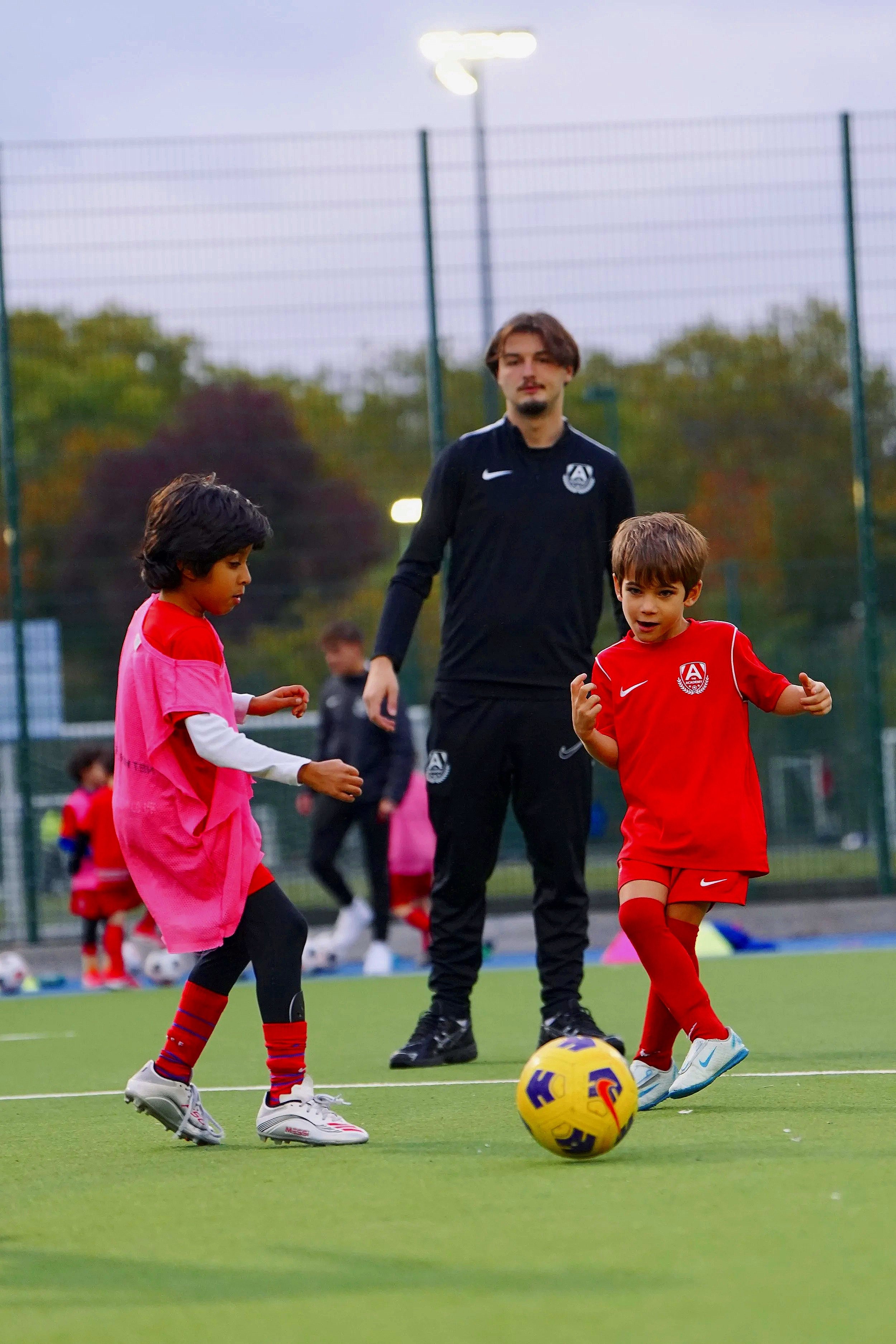 Two children playing soccer on a field, one in a pink jersey and the other in a red jersey, with an adult coach or referee watching in the background.