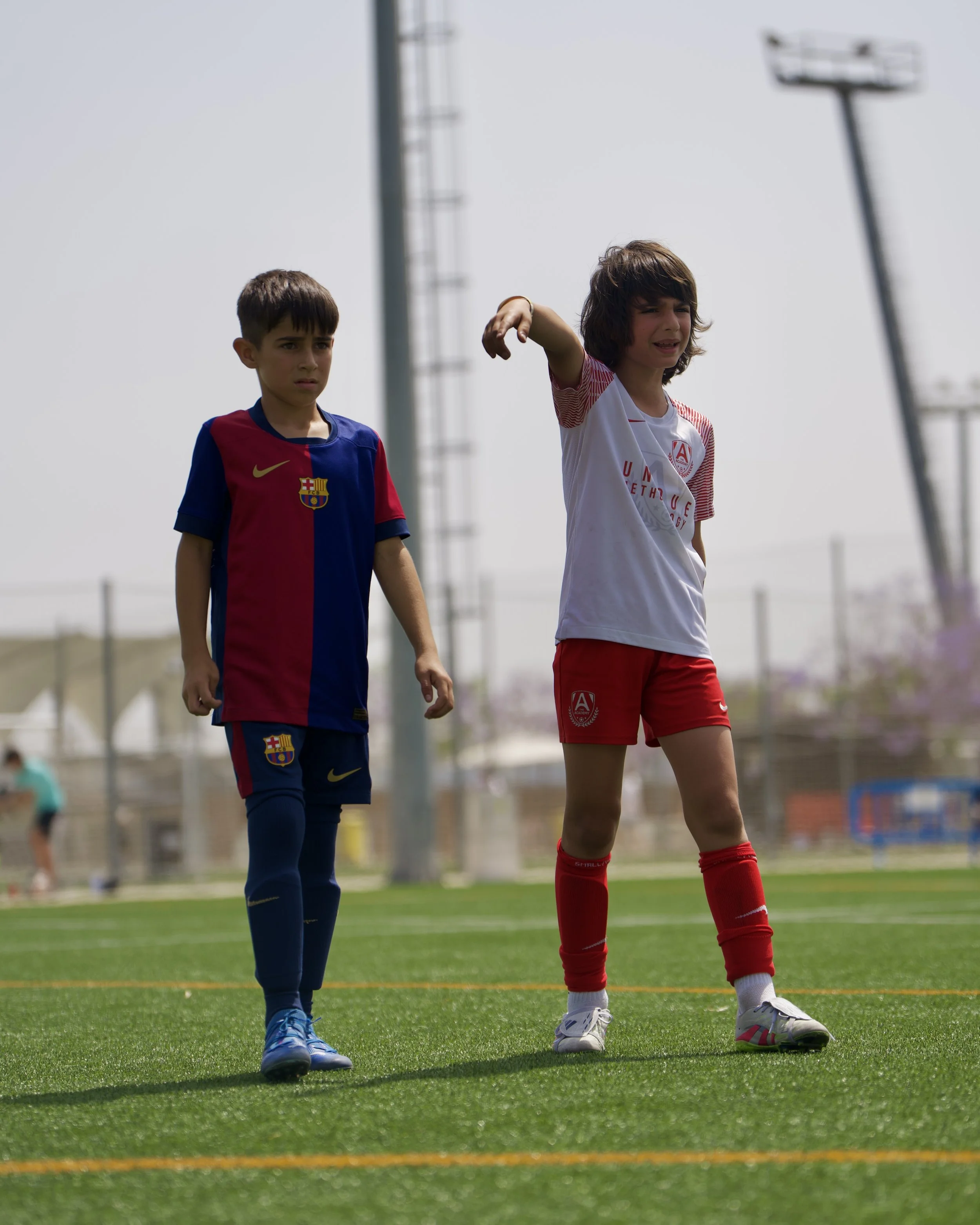Two children, a boy and a girl, standing on a soccer field. The boy wears a FC Barcelona jersey and soccer shoes, looking serious. The girl wears a white and red soccer uniform and appears to be in discomfort or upset, with her arm raised and an expression of distress.