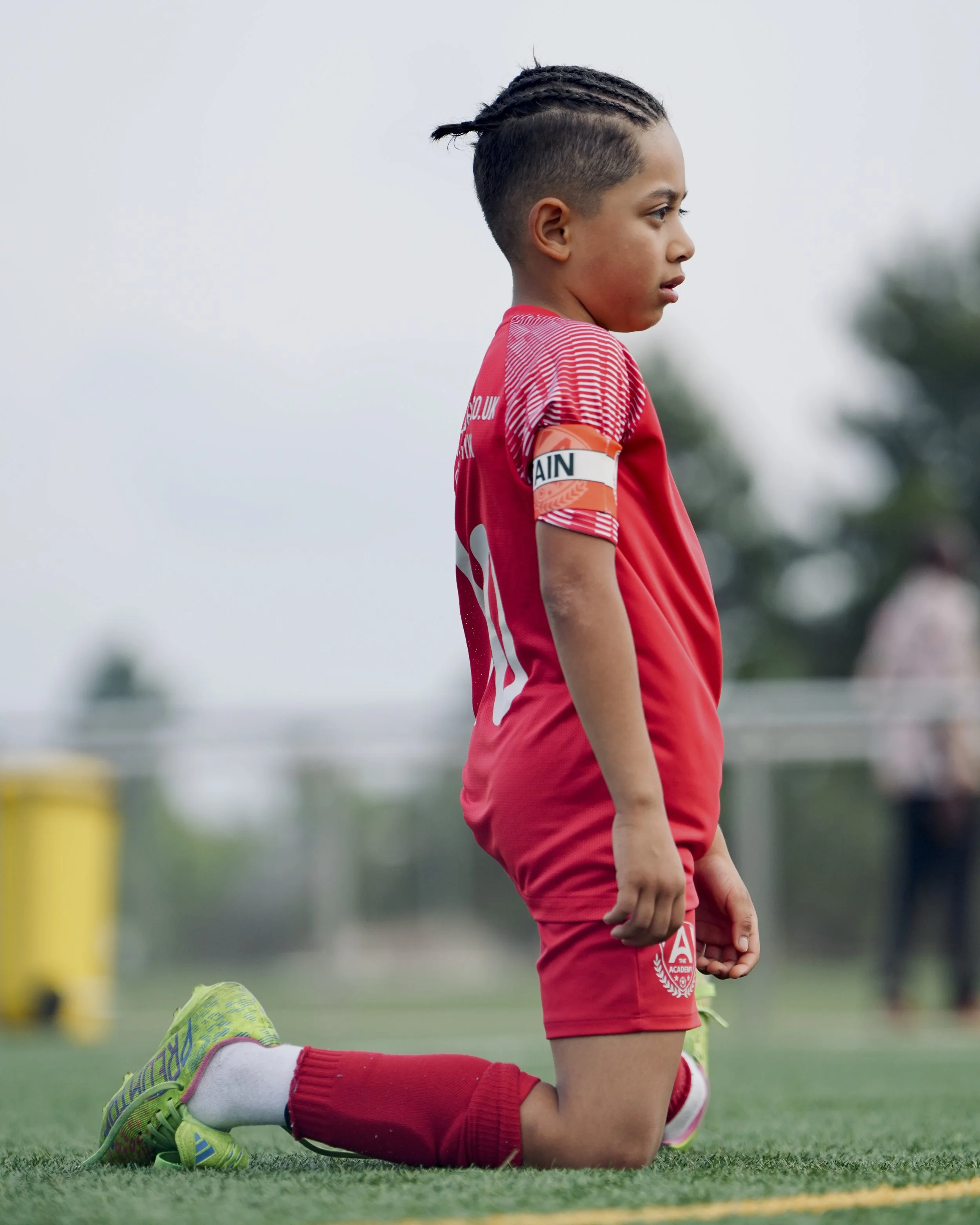 Young soccer player kneeling on grass field, wearing red uniform, with a captain's armband on his arm, looking focused.