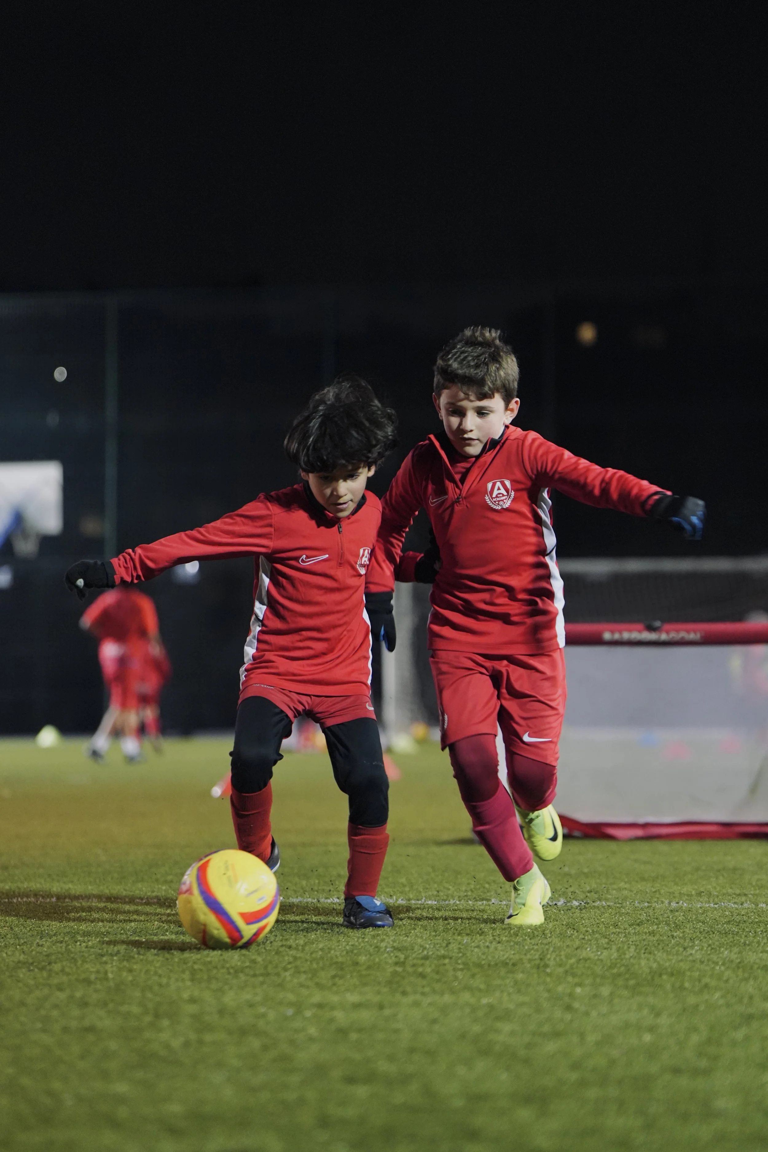 Two young boys in red soccer uniforms practicing on a field at night, playing with a yellow soccer ball.