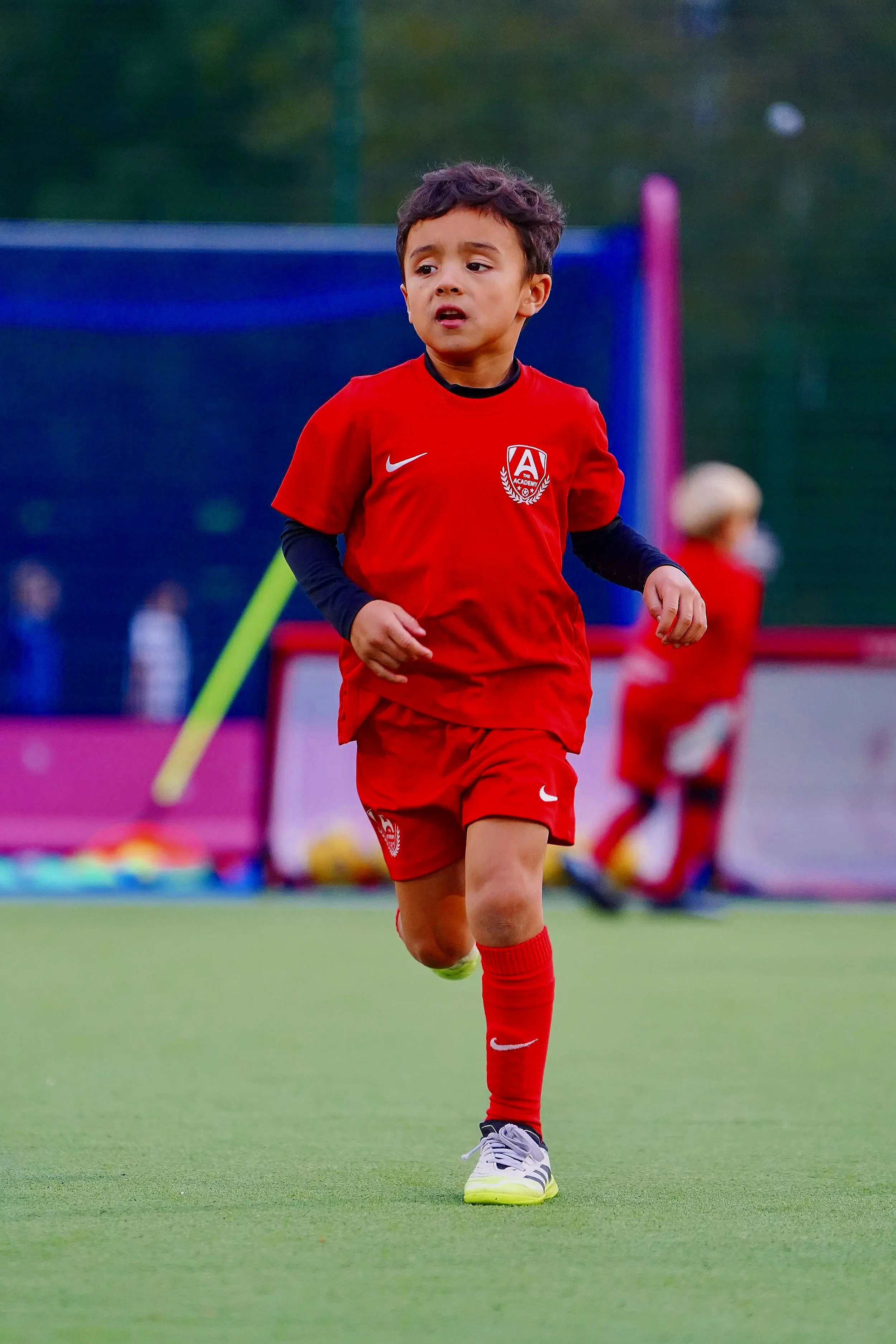 Young boy playing soccer on a field, wearing a red sports uniform with a logo on the chest, black sleeves underneath, red shorts, and soccer shoes, with trees and other children in the background.