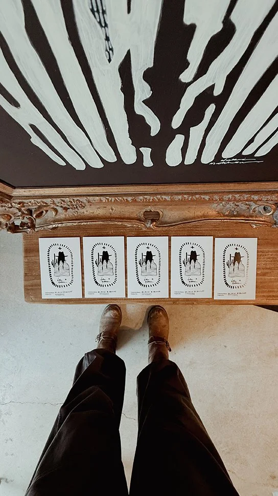 A top-down view of a person standing on a concrete floor, looking at 5 authenticated stamped cards for artwork on a wooden surface with an ornate mirror frame.