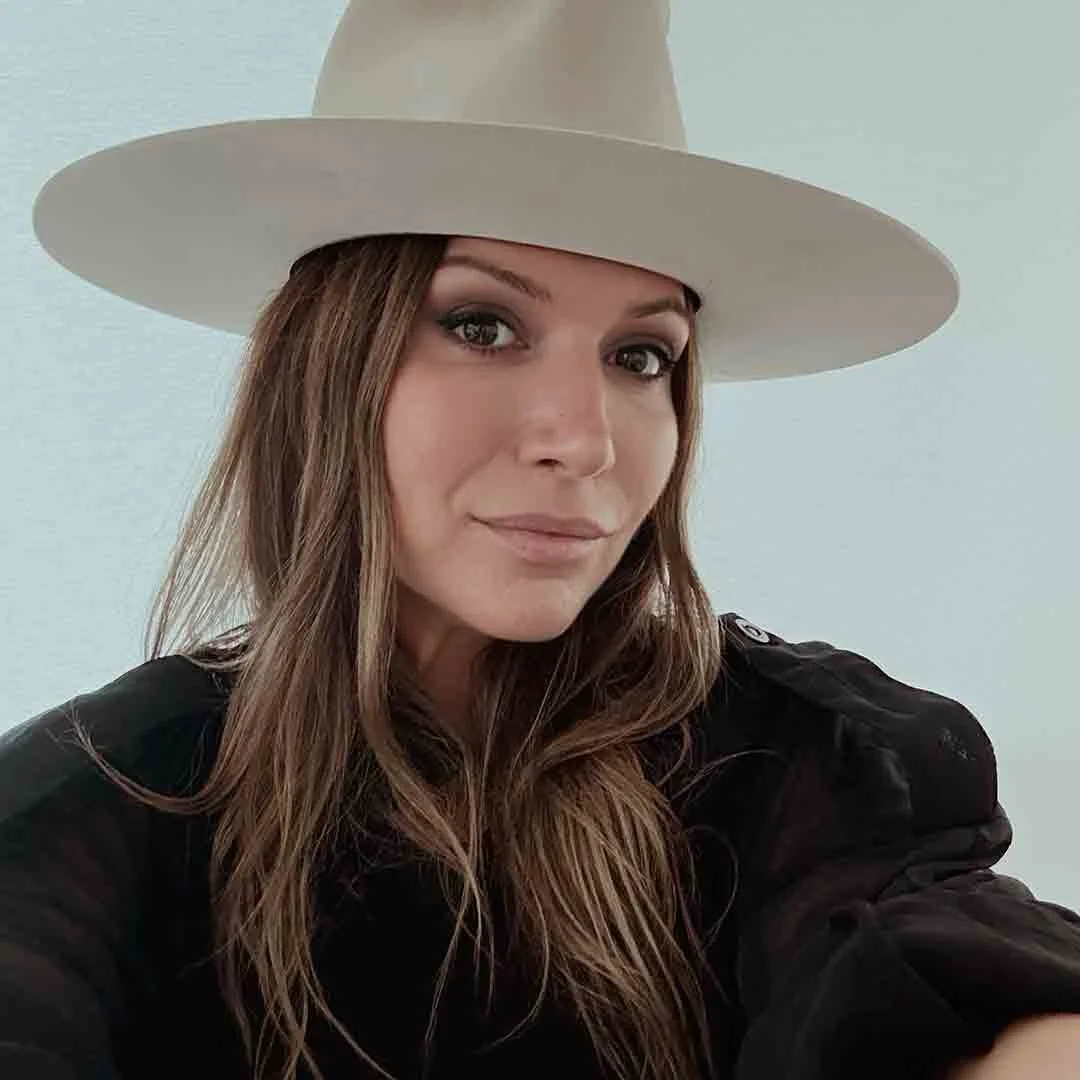 A woman with long brown hair wearing a wide-brimmed white hat and a black top, looking at the camera with a slight smile.