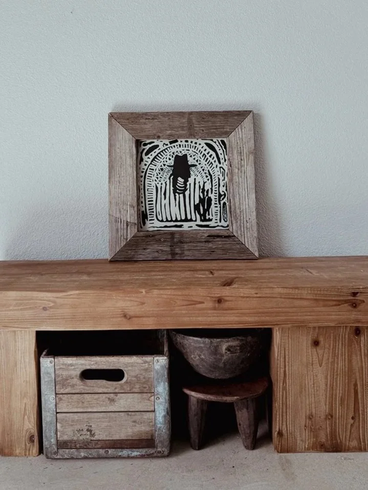 A framed black and white abstract artwork rests on a wooden shelf against a light wall, with a rustic wooden box and a small wooden stool with a bowl on top below.