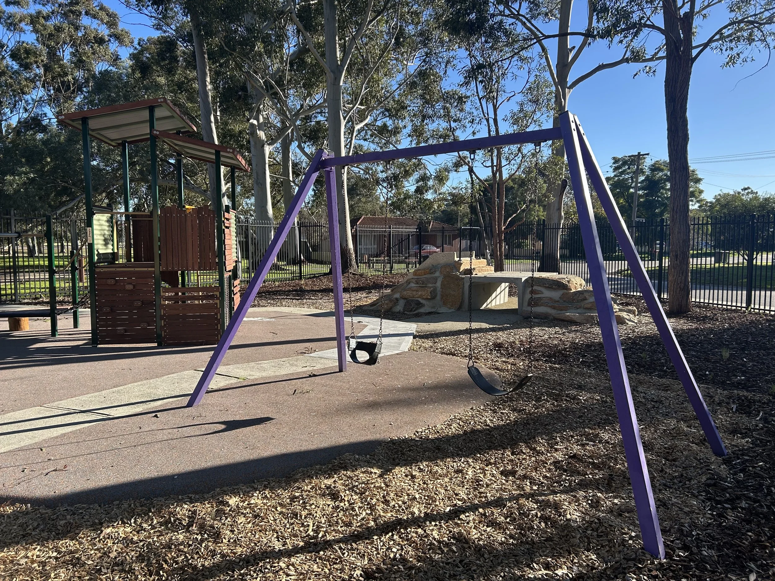 Empty playground with a purple swing set, a wooden play structure, and a rock climbing wall, surrounded by trees and a black metal fence.