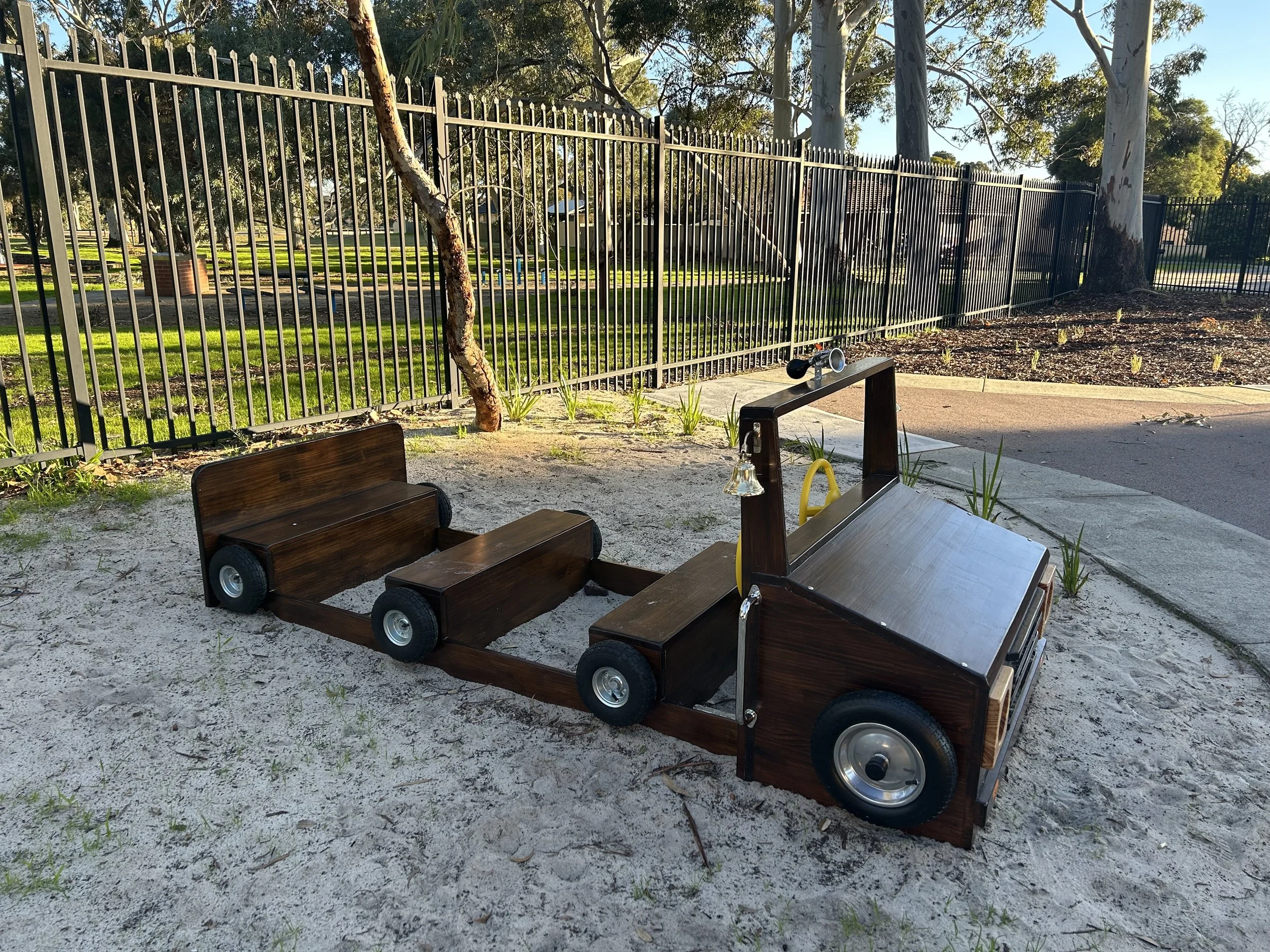 A wooden toy truck played on sandbox with grass and trees in background, black metal fence in park area.