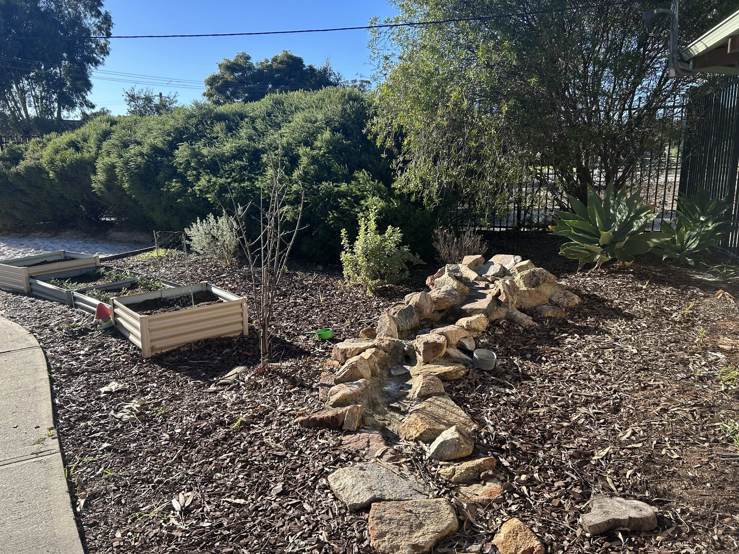 A garden with a stone-lined dry creek bed, small raised garden beds, bushes, and succulents along a fence and sidewalk under a blue sky.