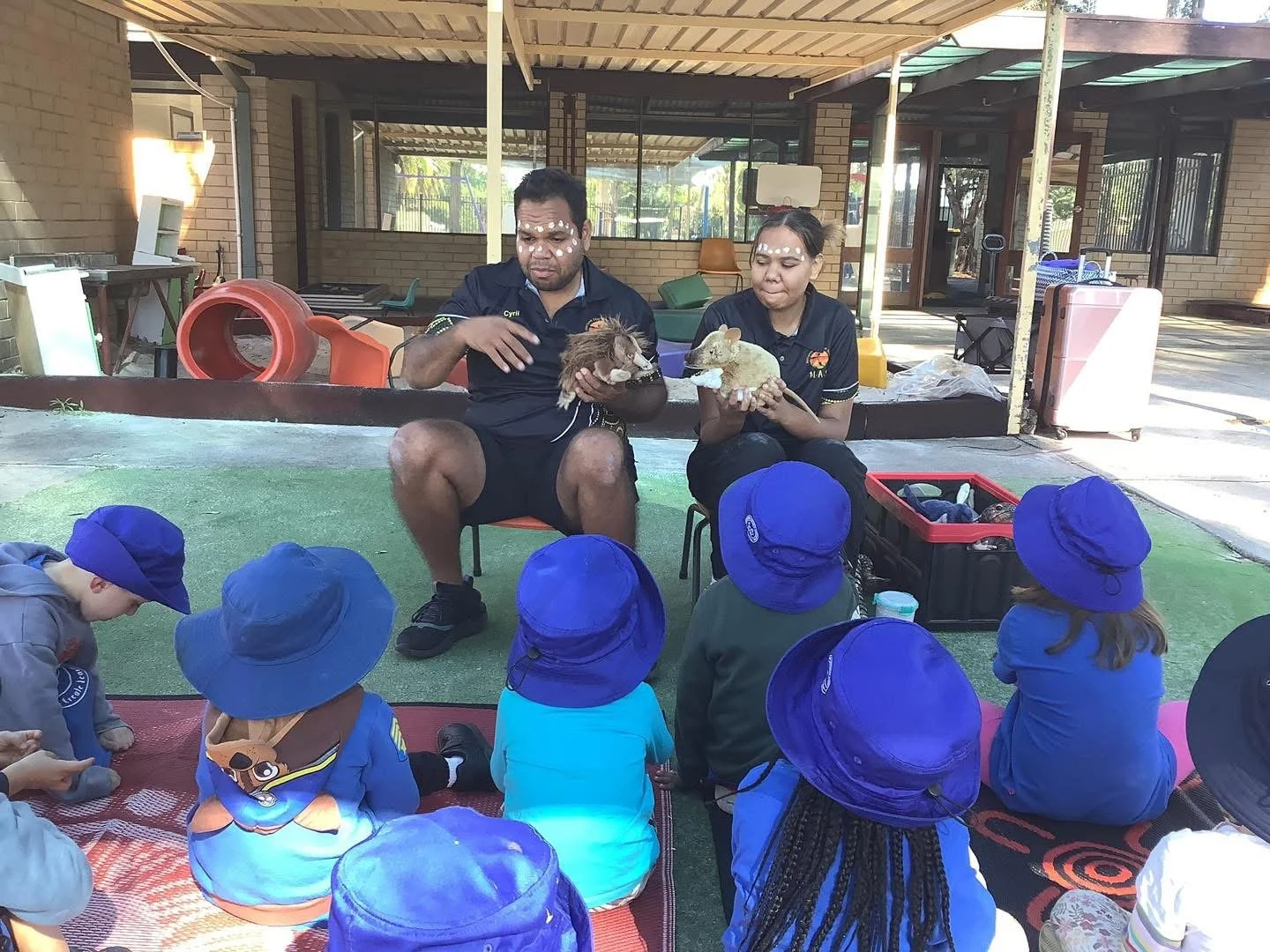 Two animal handlers demonstrating animals to a group of young children, including a man holding a small animal and a woman holding a different animal, with children wearing blue hats sitting on mats in an outdoor educational setting.
