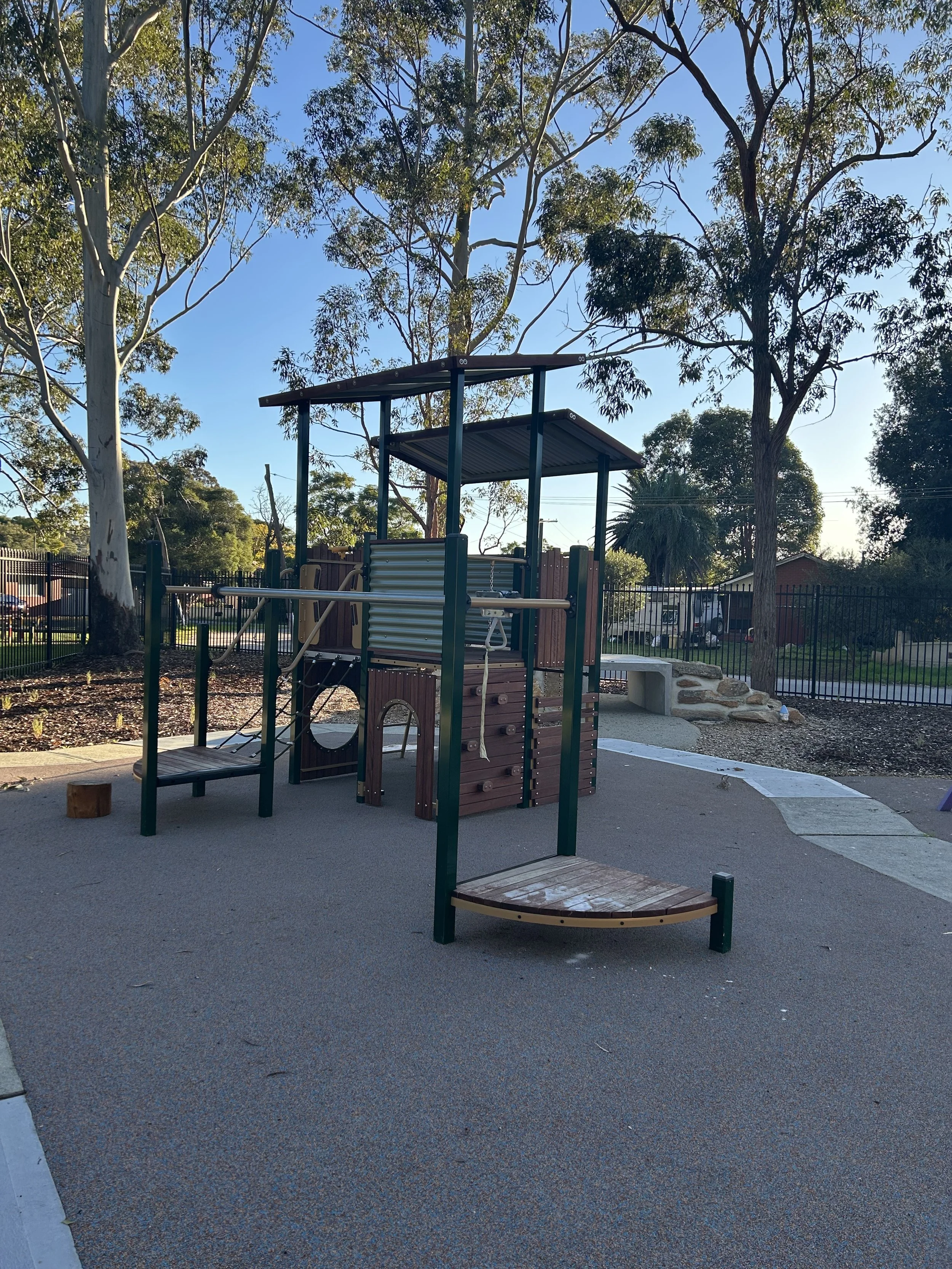 Playground with climbing structure, slide, and wooden stepping stones under a clear blue sky, surrounded by trees.