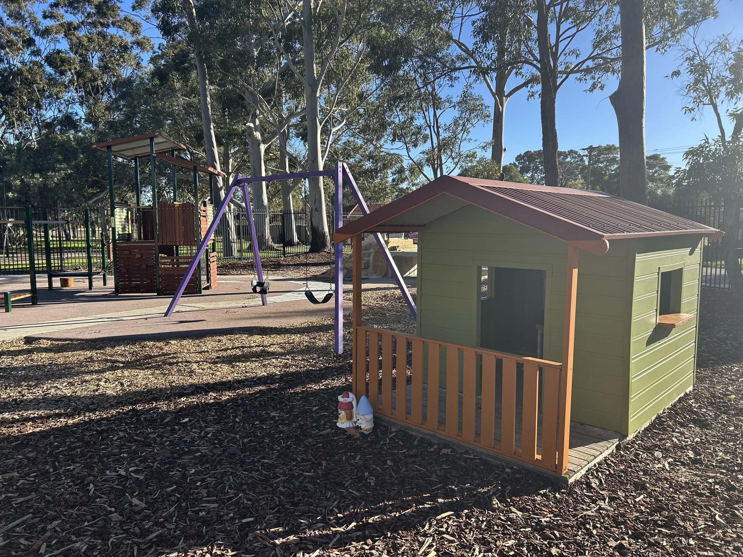 Children's playground with a small playhouse, swings, and climbing structures surrounded by tall trees on a sunny day.