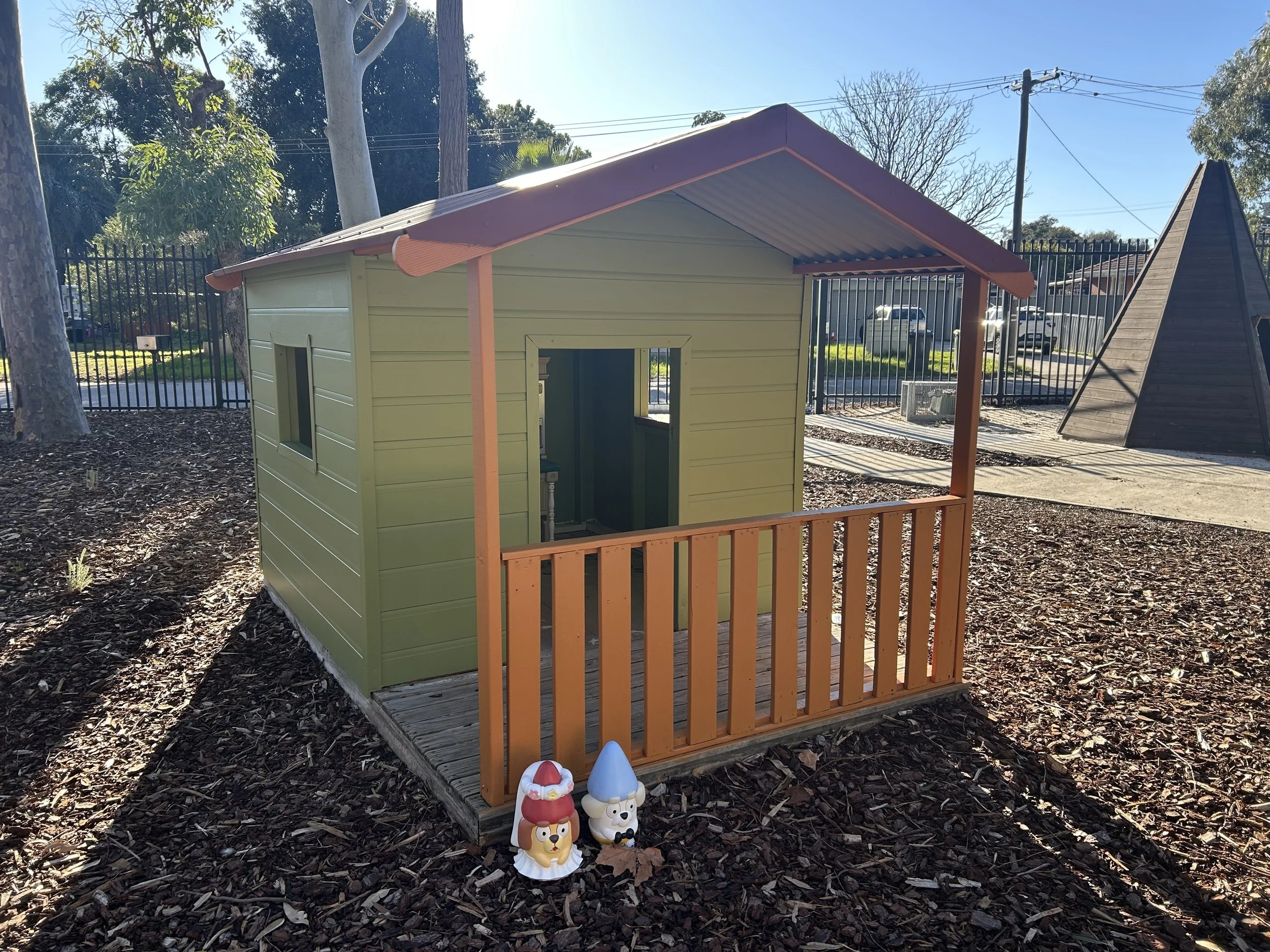 Small playhouse painted in green with orange railings and a brown sloped roof, located in a playground with mulch ground, a tree, and a wooden pyramid structure nearby.
