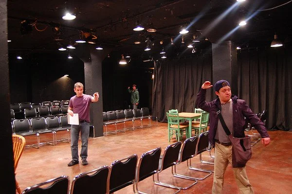 Two men in a black-box theater with rows of black chairs, some green chairs, and a black curtain, practicing or rehearsing.