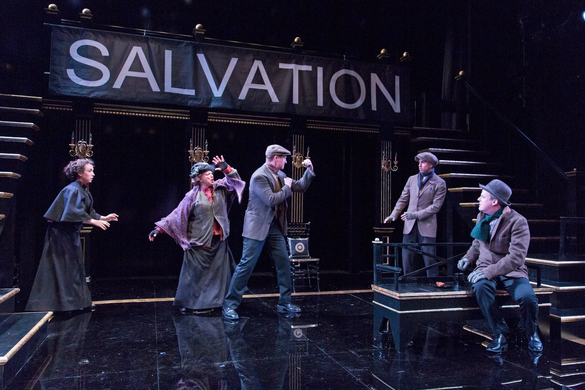 Theatrical performance with five actors on stage, a large "SALVATION" banner in the background, dark stage setting, actors in period costumes, some gesturing dramatically.