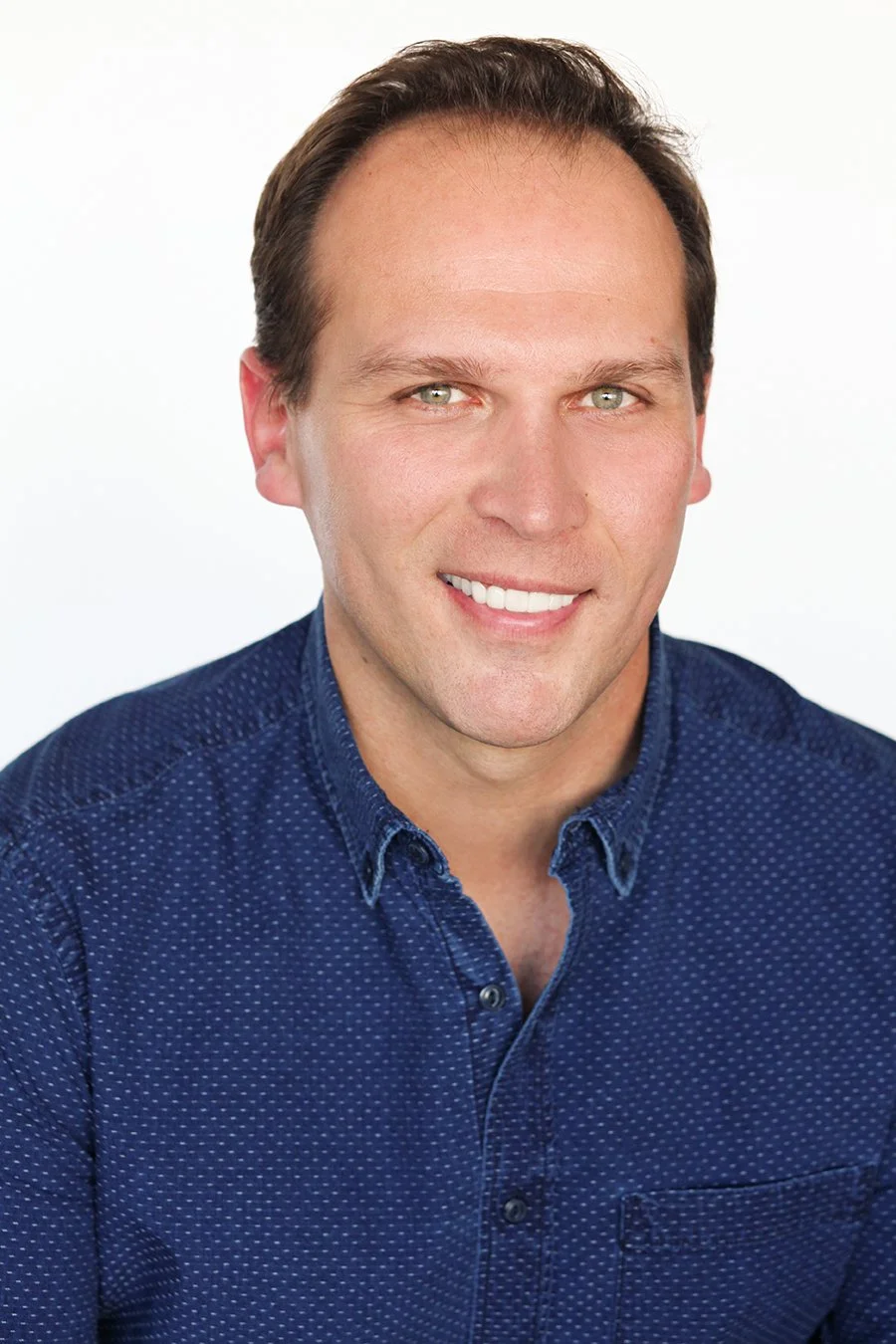 Headshot of a smiling man with short brown hair, light skin, wearing a blue button-up shirt with small white dots.