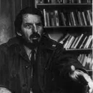 A man with dark hair and a beard, wearing a dark shirt or jacket, sitting in front of a bookshelf filled with books.