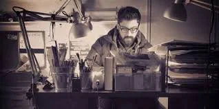 A man with glasses working at a cluttered desk with a laptop, desk lamp, and stacks of papers in a dimly lit office.
