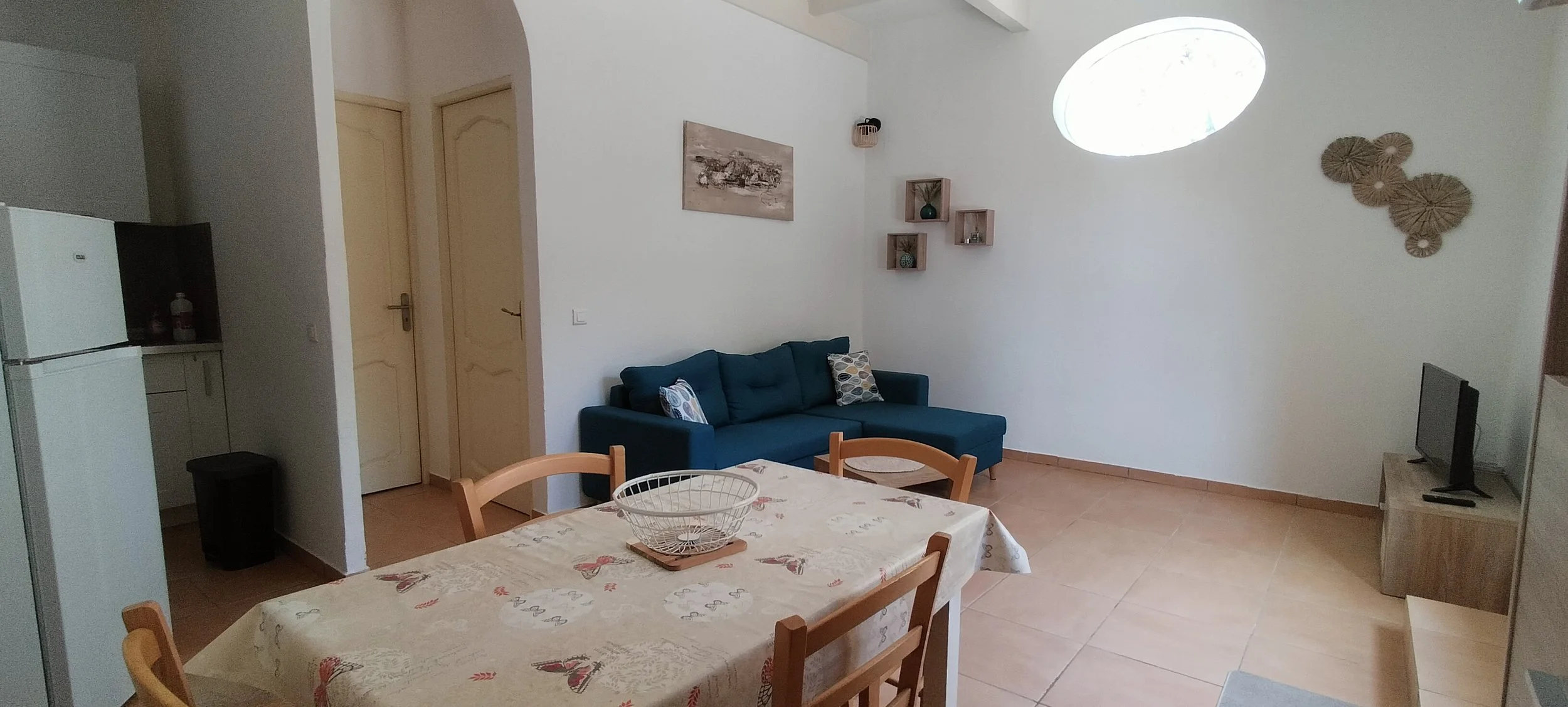 Salon avec canapé bleu, table à manger avec nappe, petit téléviseur, décorations murales en osier, étagères avec vases, et une lucarne au plafond permettant la lumière naturelle.