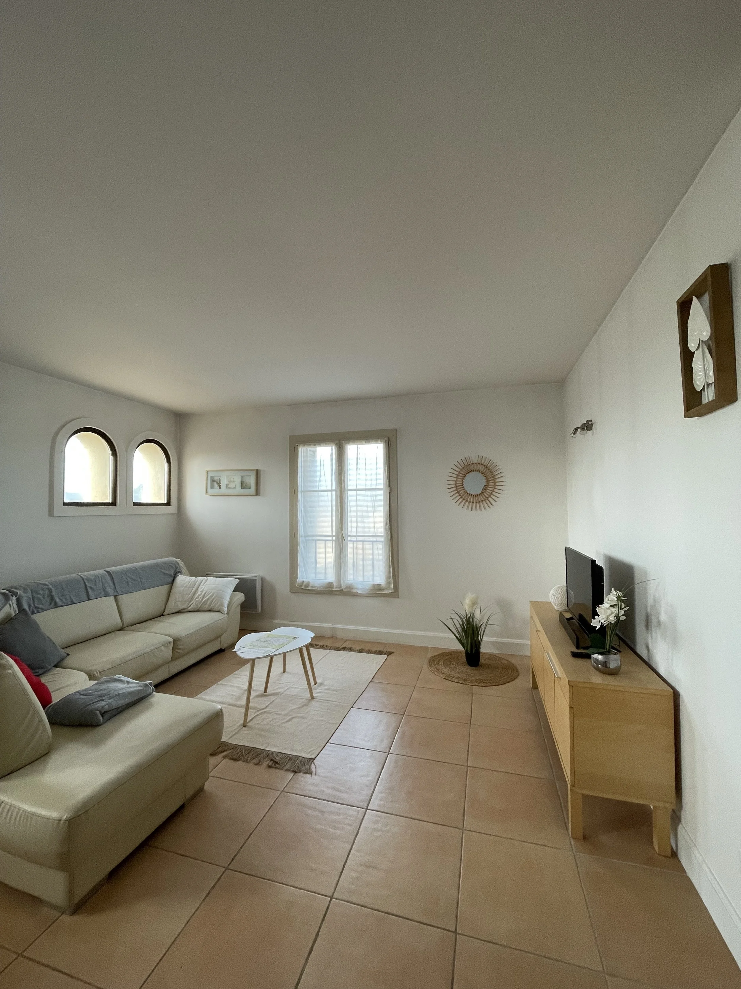 Salon lumineux avec un canapé beige, table basse, télévision sur un meuble en bois, plantes décoratives, et fenêtres avec rideaux blancs.