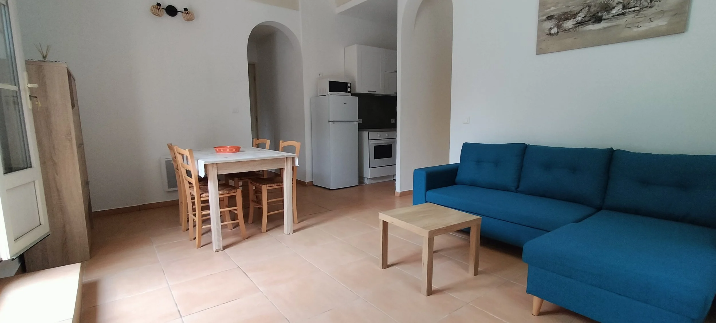 Salon avec un canapé bleu, une table en bois, une cuisine avec réfrigérateur, four, micro-ondes, et une salle à manger avec quatre chaises en bois.