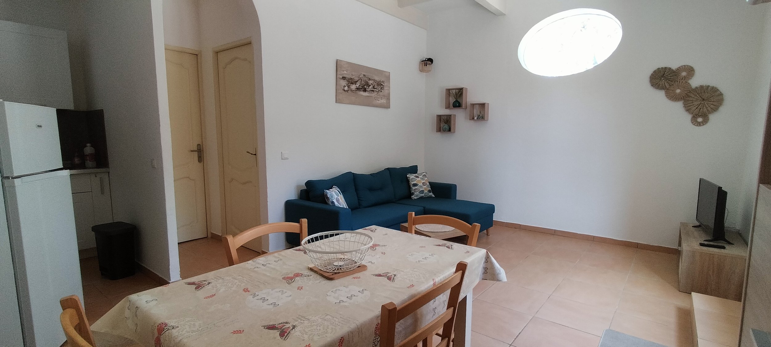 Salon avec canapé bleu, table à manger avec nappe, petite télévision sur meuble, décorations murales en rotin, fenêtres ovales. Cuisine visible avec réfrigérateur et placard.