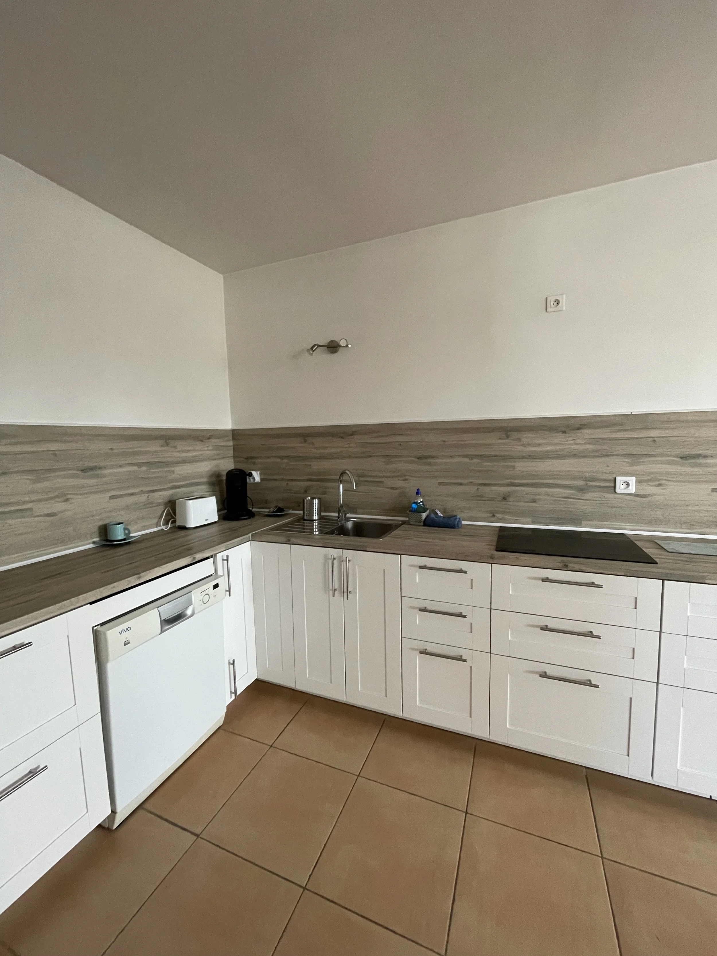 Cuisine moderne avec plans de travail en bois clair, meubles en blanc, évier en acier inoxydable, petite électroménager noir et blanc, et des tasses bleues. Sol en carreaux beige.