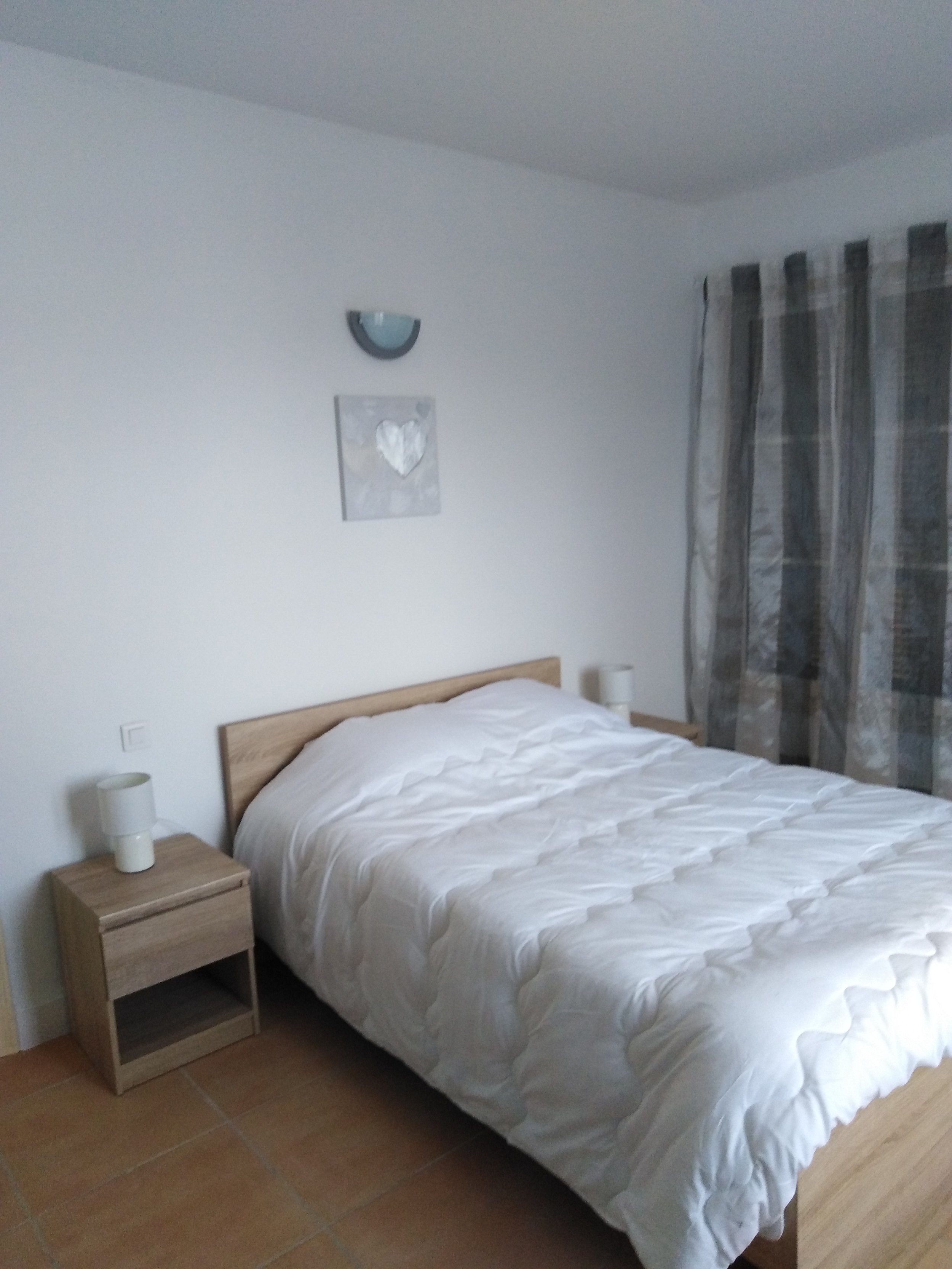 Chambre simple avec lit en bois, deux lampes de chevet, rideaux gris et tableau de cœur blanc sur le mur.