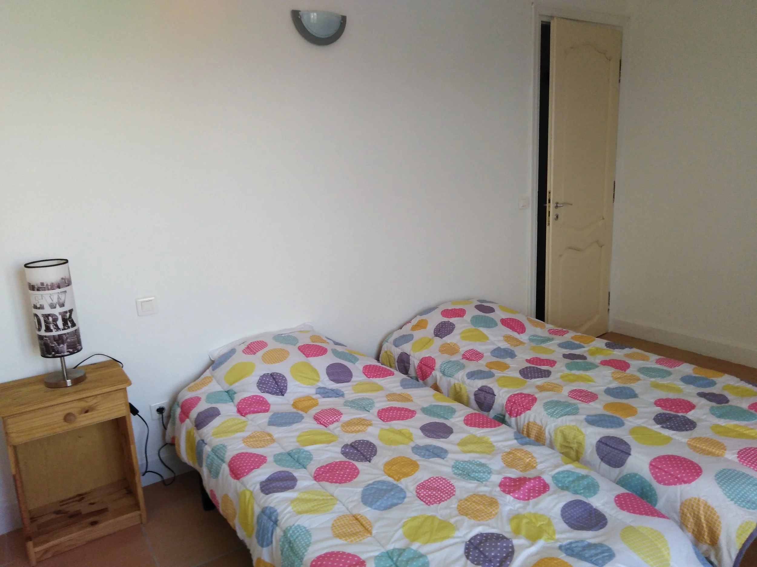 Chambre avec deux lits simples avec couverture à pois colorés, une lampe de chevet vintage, et une porte entrouverte.