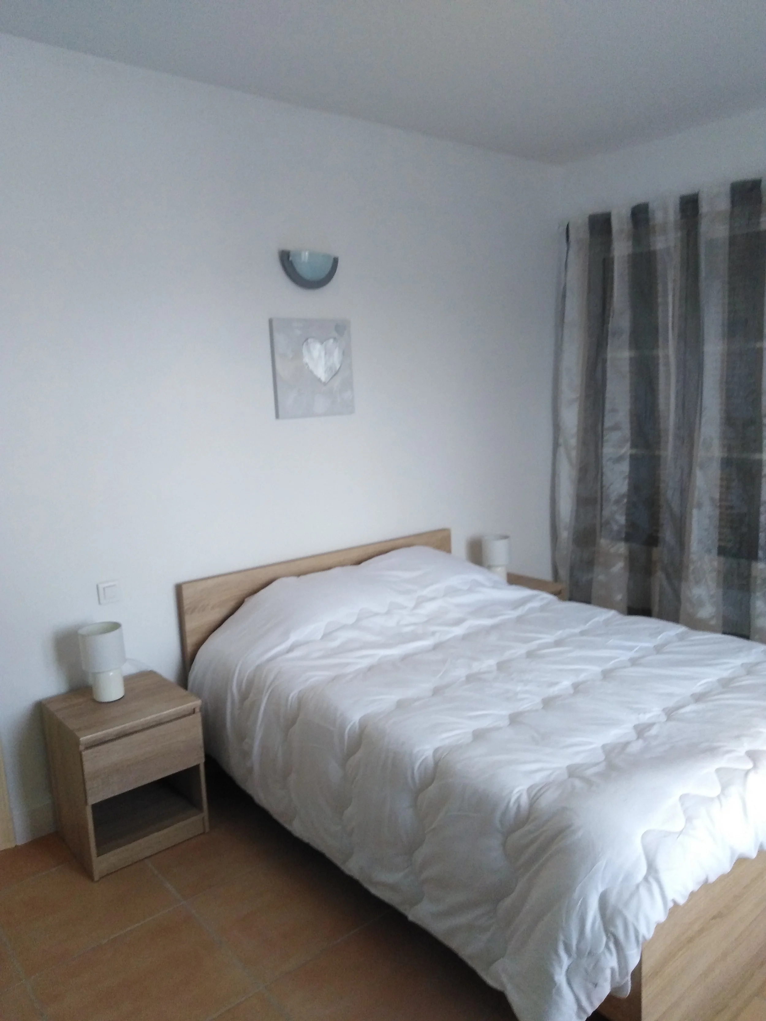 Chambre à coucher simple avec un lit en bois, une tête de lit en bois clair, et une literie blanche. Il y a deux tables de chevet avec une lampe chacune, un tableau avec un cœur, et un luminaire mural. Un rideau à motifs terne couvre une fenêtre.
