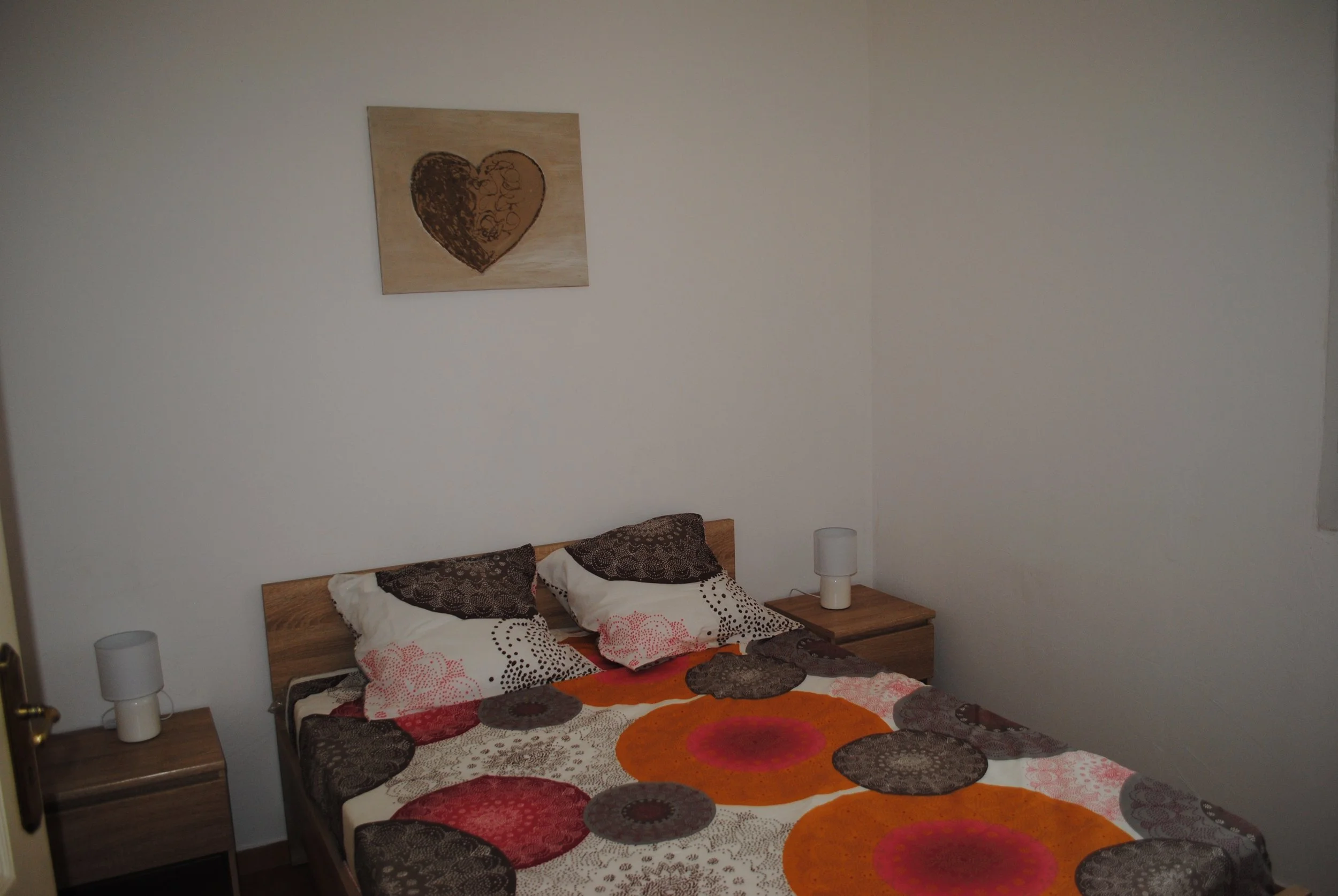 Chambre simple avec un lit double, deux tables de nuit avec lampes et un tableau de cœur au mur.