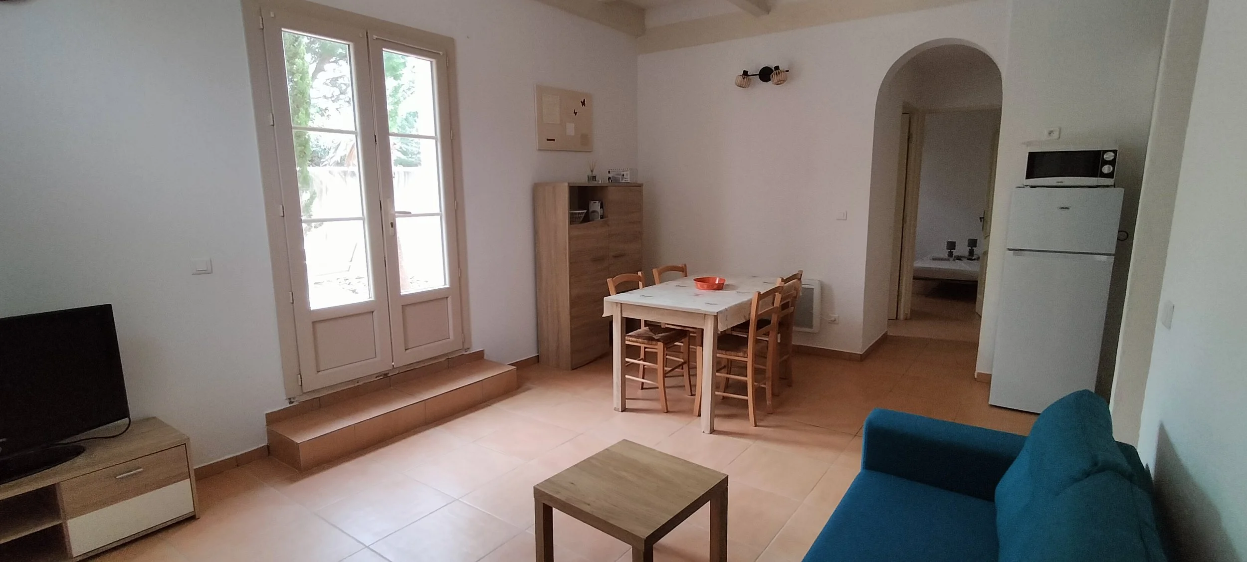 Intérieur d'un salon avec une télévision, un canapé bleu, une table basse, une table à manger avec des chaises en bois, un réfrigérateur avec un micro-ondes dessus, une porte-fenêtre donnant sur l'extérieur, et un espace cuisine visible à travers une