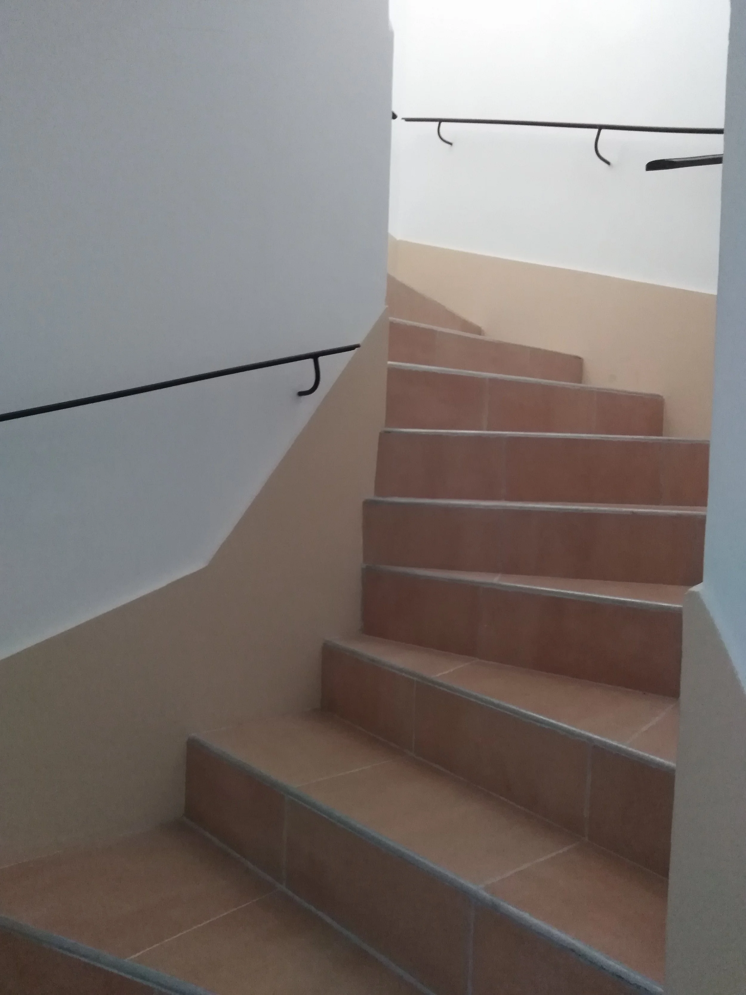 Escalier en terre cuite avec main courante noire, vue de bas en haut, dans un bâtiment coloré beige et blanc.