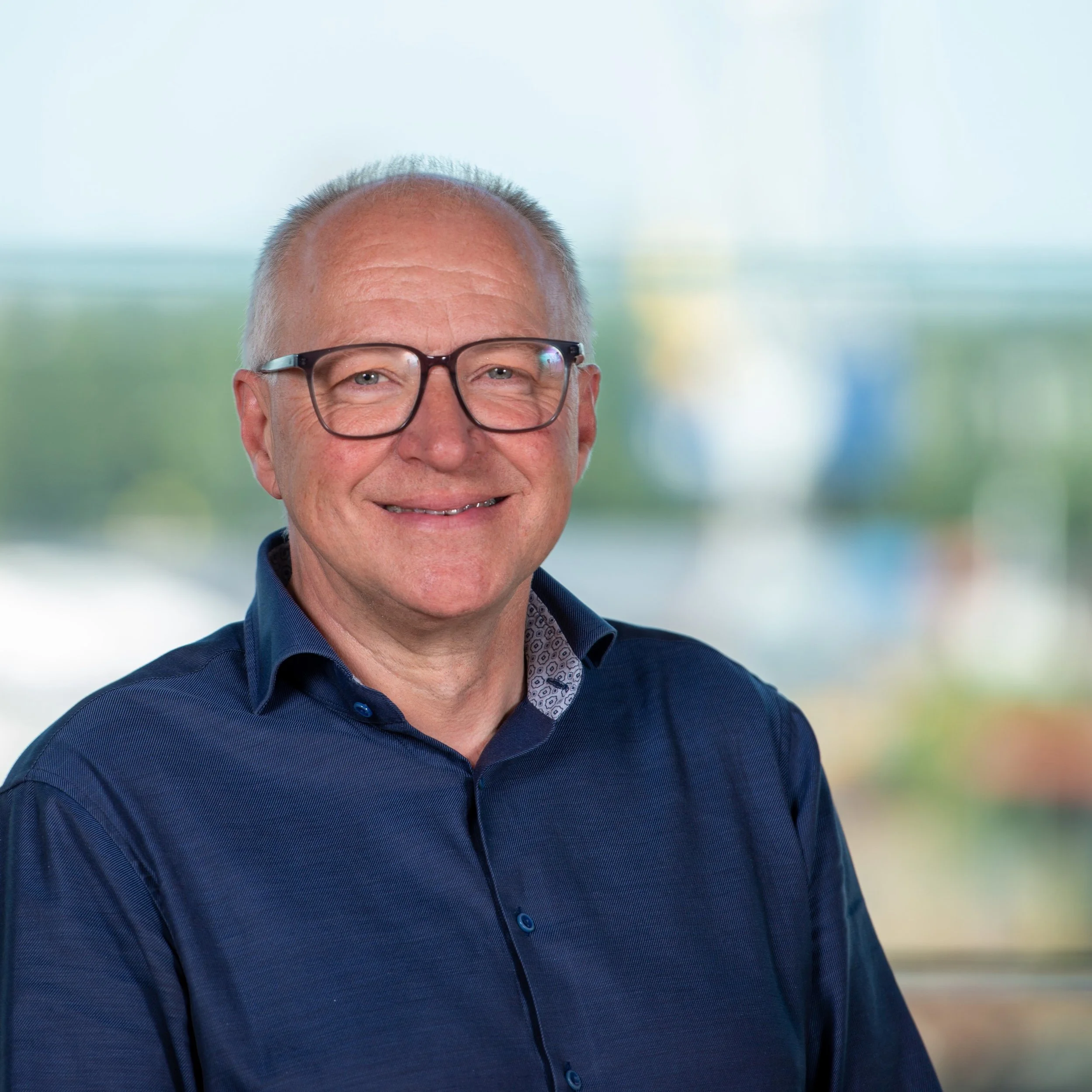 Portretfoto van een lachende man met kort grijs haar, grote bril en donkerblauw overhemd, op achtergrond van een raam met uitzicht op buiten.