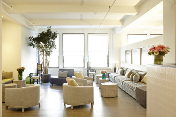 Beautifully furnished bright living room with large windows, beige chairs, a long striped sofa with cushions, potted plants, and fresh flowers.