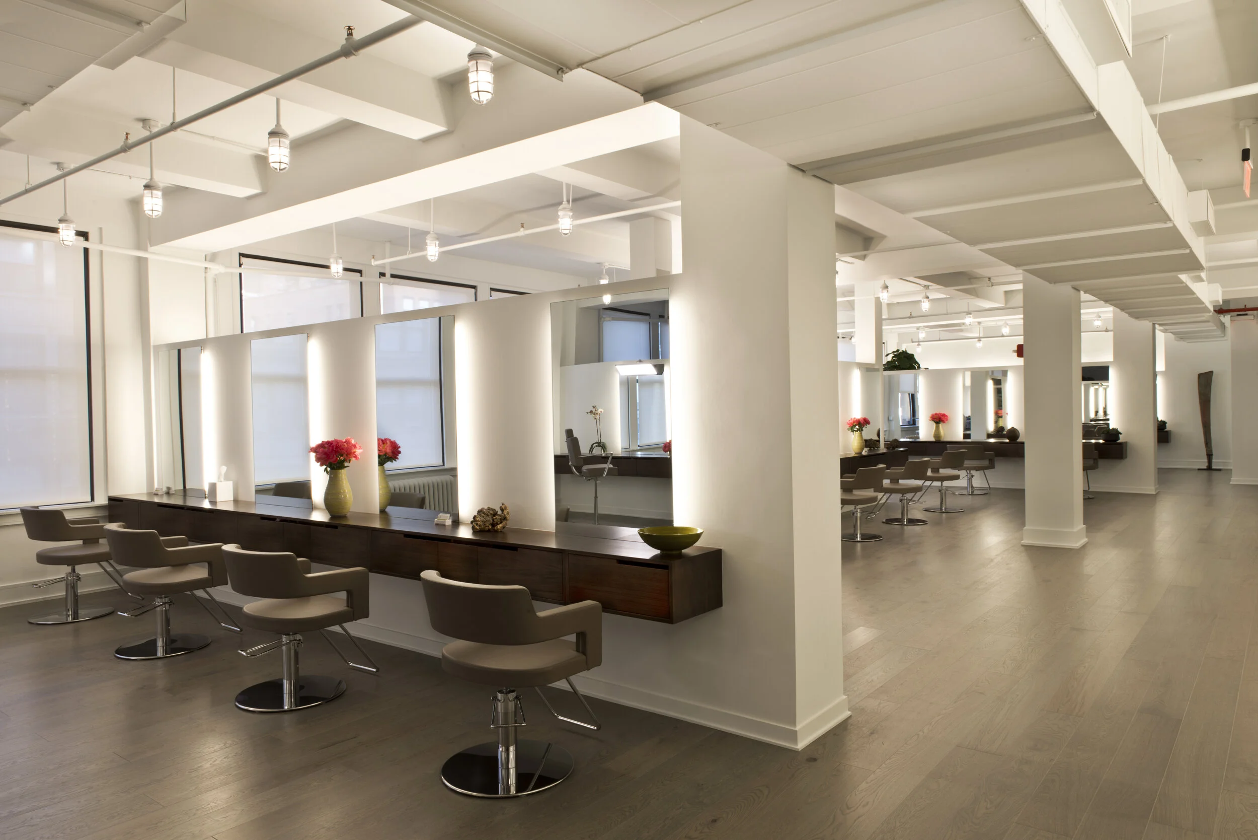 Modern hair salon with styling stations, chairs, mirrors, large windows, and decorative vases with flowers.