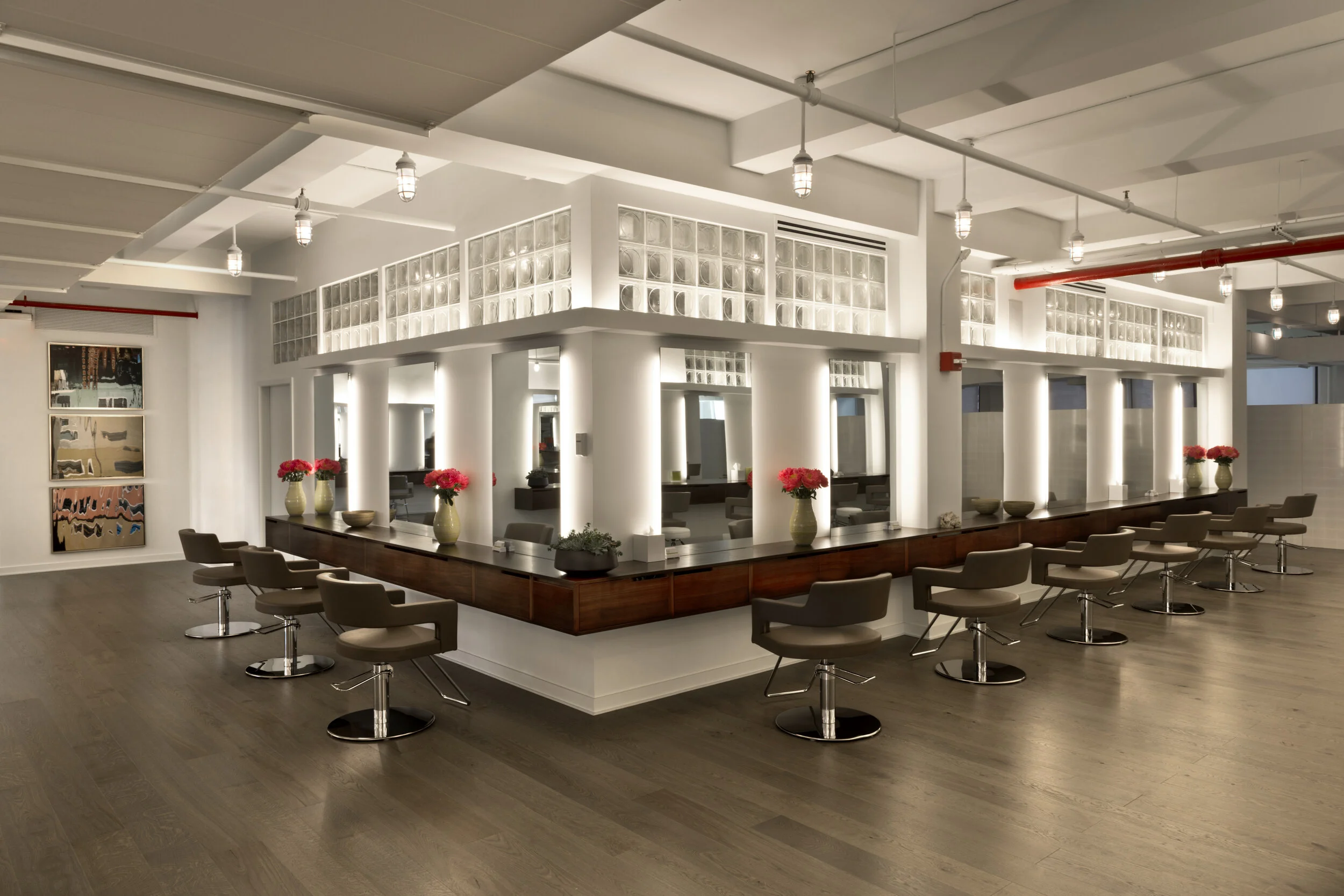 Modern hair salon interior with a large, circular station wrapped in a wooden countertop, surrounded by adjustable height salon chairs with beige cushions. The station has decorative vases with pink flowers. The salon features white walls, ample ligh