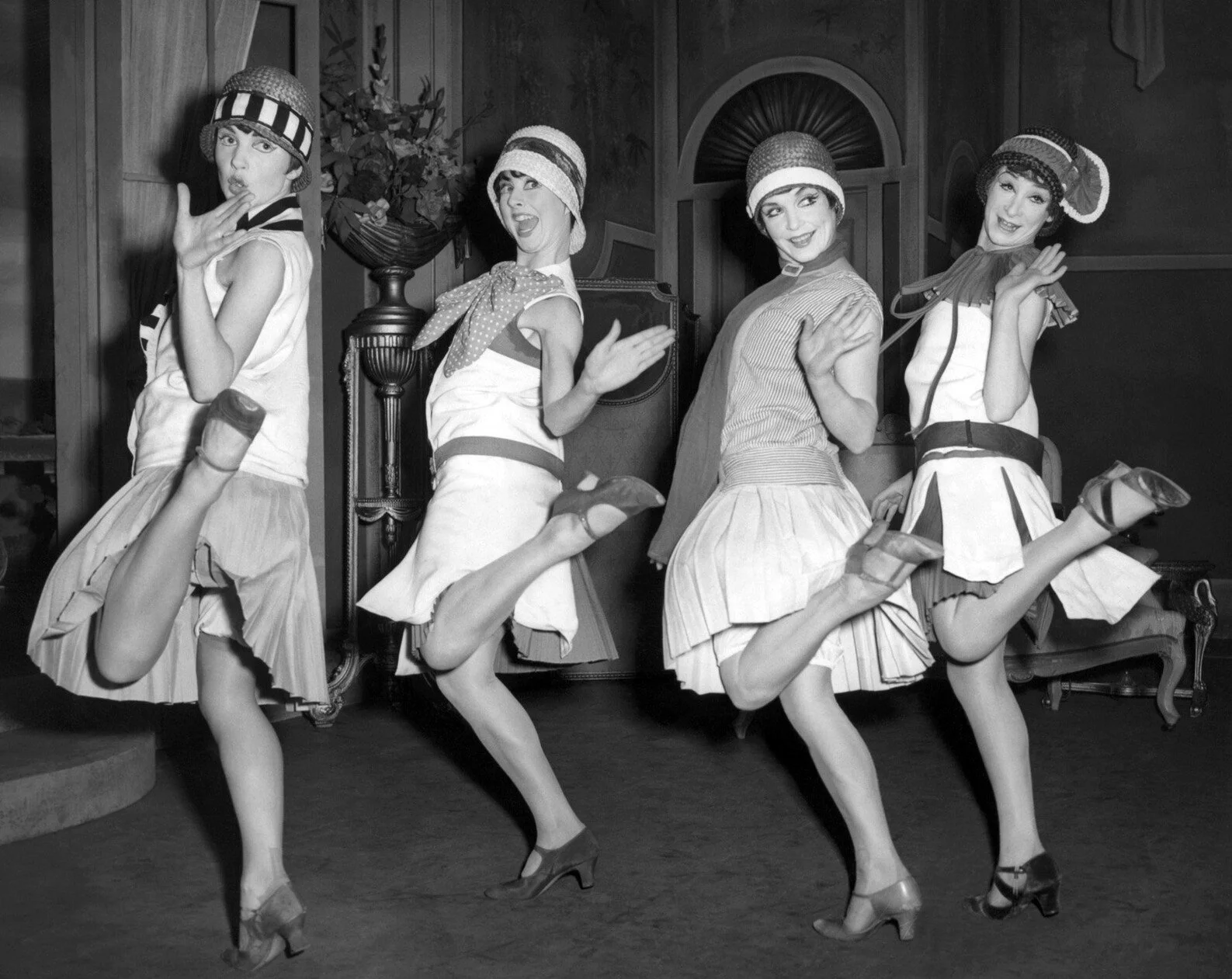 Four women in 1920s flapper dresses and cloche hats performing a dance routine, kicking their legs sideways in a vintage setting.