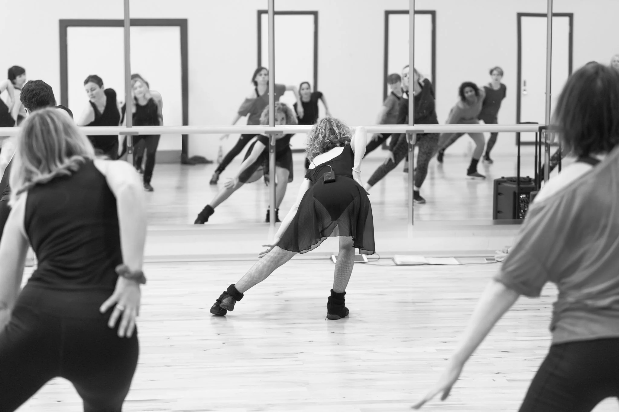 Black and white image of a dance class with people stretching, mirrored wall.