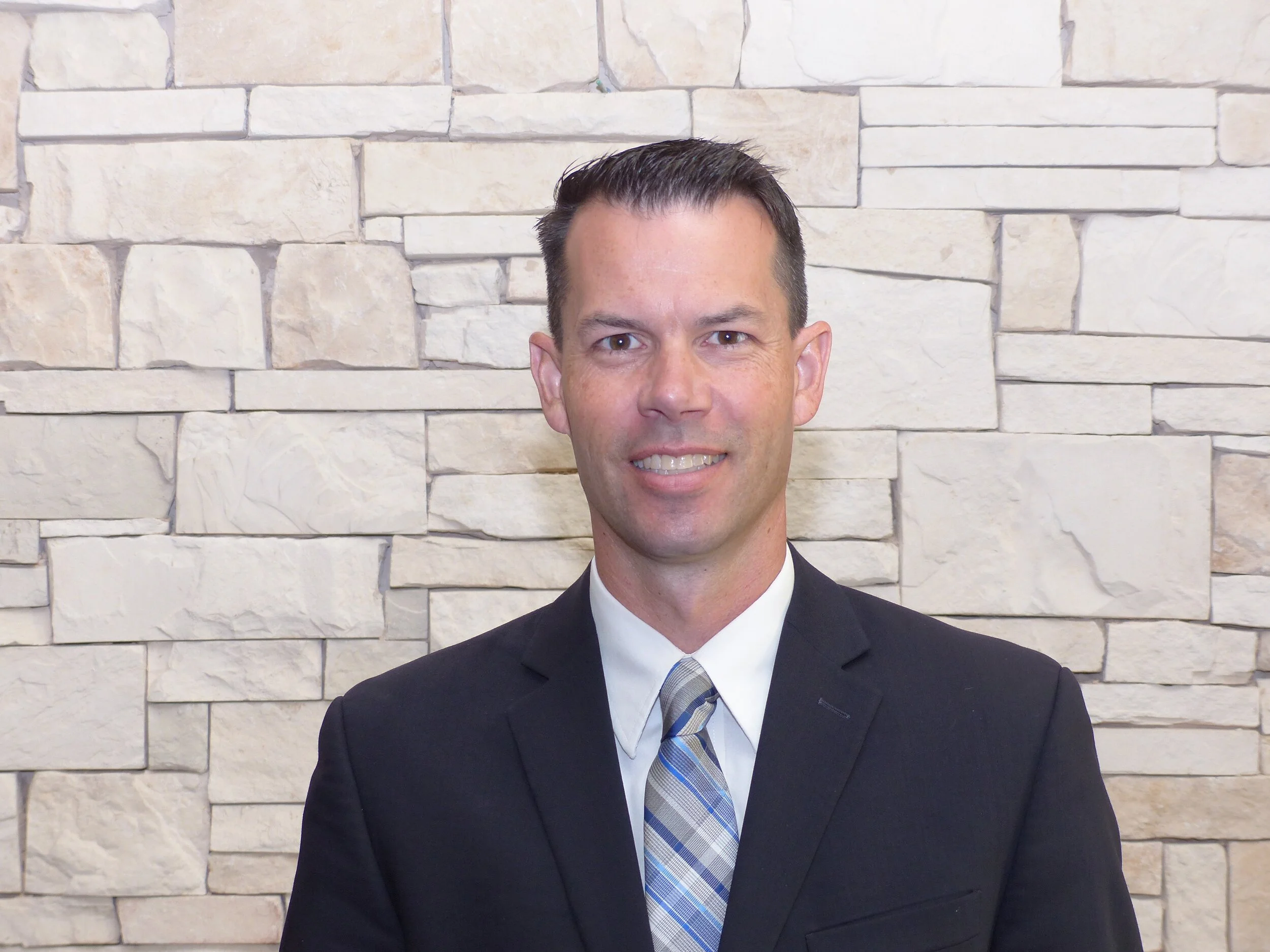 Portrait of a man in a white shirt, striped tie, and dark suit jacket standing in front of a beige stone wall.