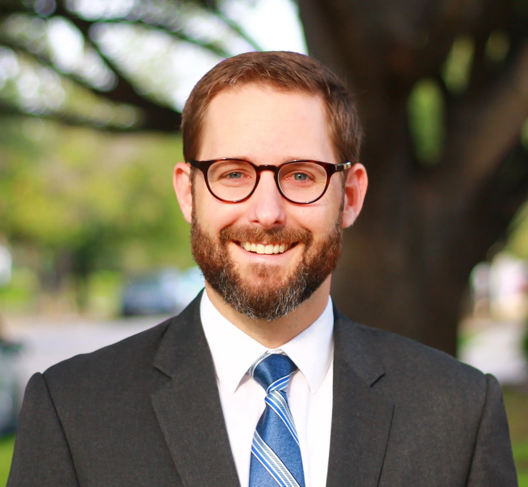 A smiling man with glasses and a beard wearing a suit and tie, outdoors in front of a tree.